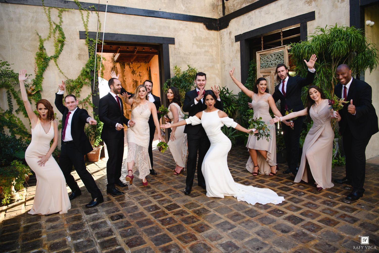 Wedding party celebrating outdoors in courtyard, bride in white gown surrounded by bridesmaids and groomsmen cheering.