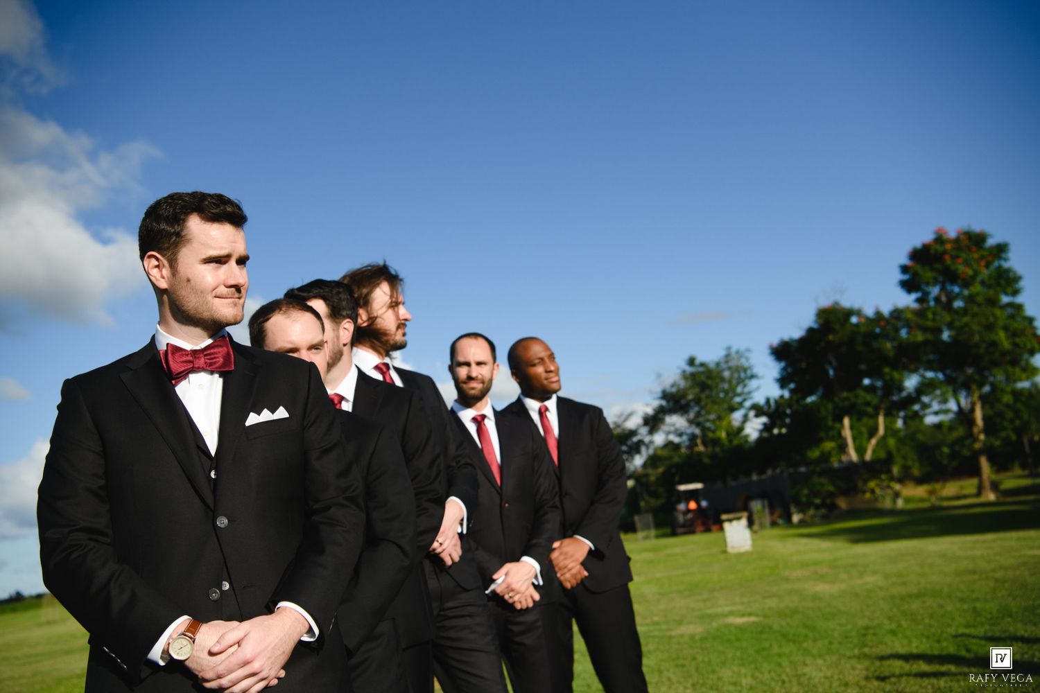 Groomsmen in black tuxedos with red bow ties stand outdoors on a sunny day.