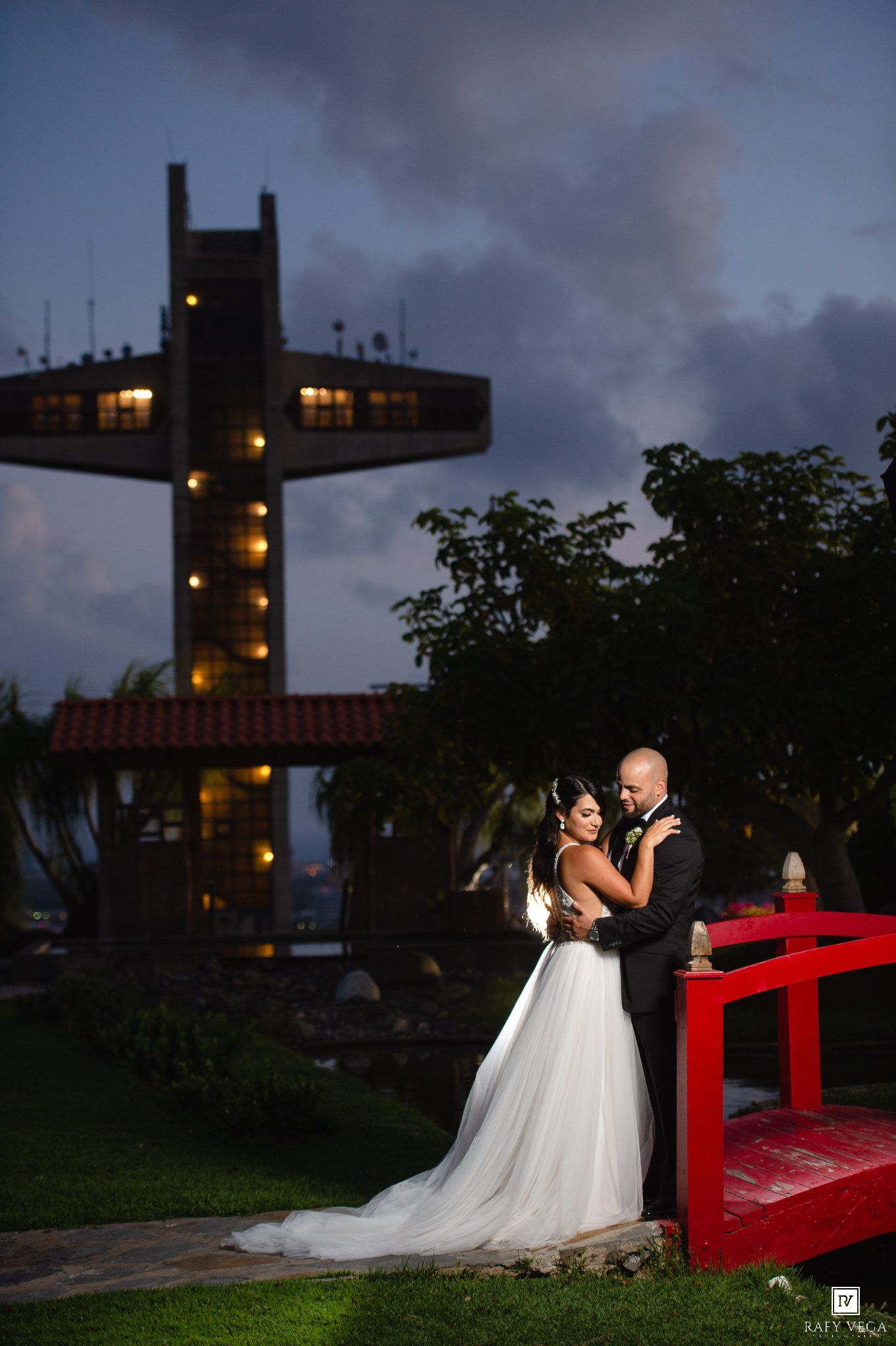 Bride and groom embrace at dusk near a red bridge with a cross tower backdrop at an elegant outdoor wedding venue.