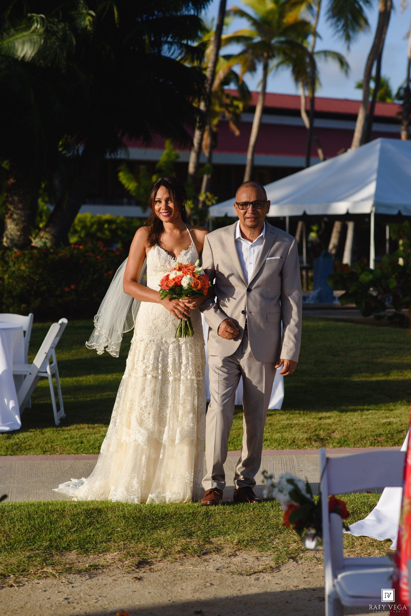 Bride in lace gown holding coral bouquet walks with escort at outdoor tropical wedding ceremony.