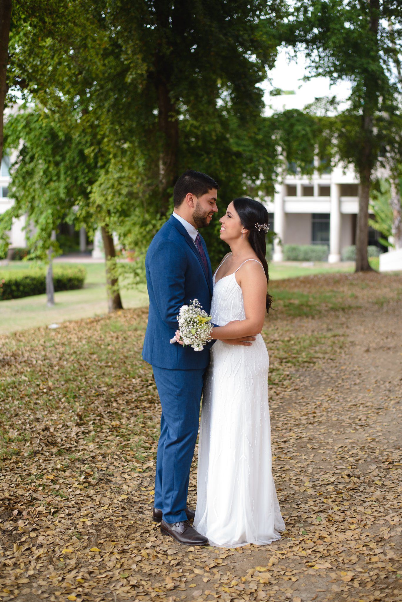 Bride and groom share a romantic moment in a park, she holds white flowers, he wears a navy suit, lush trees behind them.