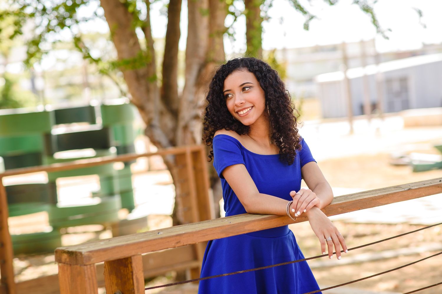 Young woman in blue dress smiling while leaning on wooden fence outdoors on sunny day.