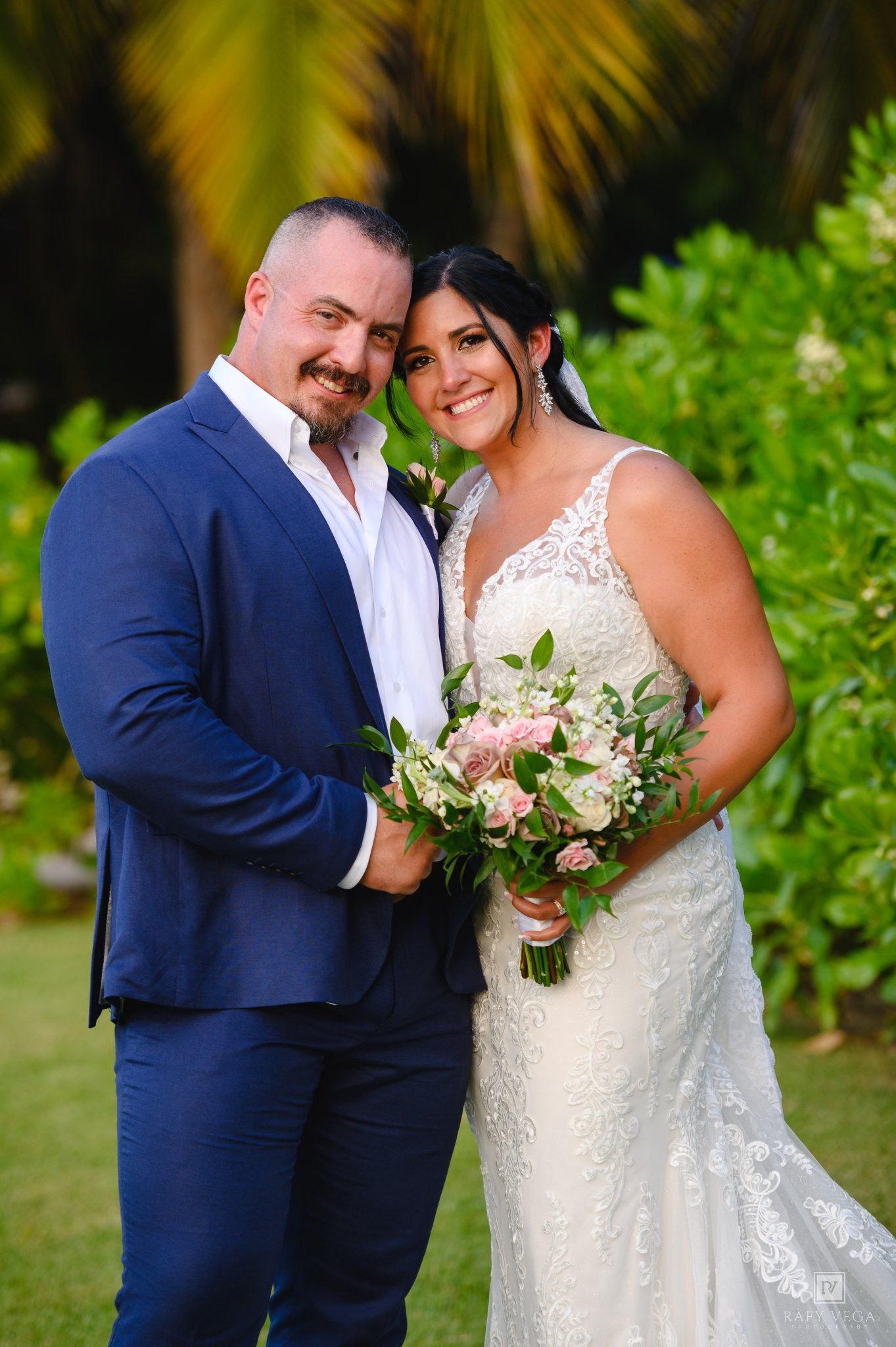 Smiling bride in lace gown holding pink bouquet poses with groom in navy suit outdoors at tropical wedding venue.