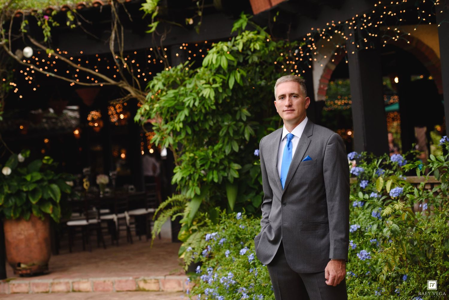 Elegant man in gray suit and blue tie standing in lush garden courtyard with string lights at formal event.