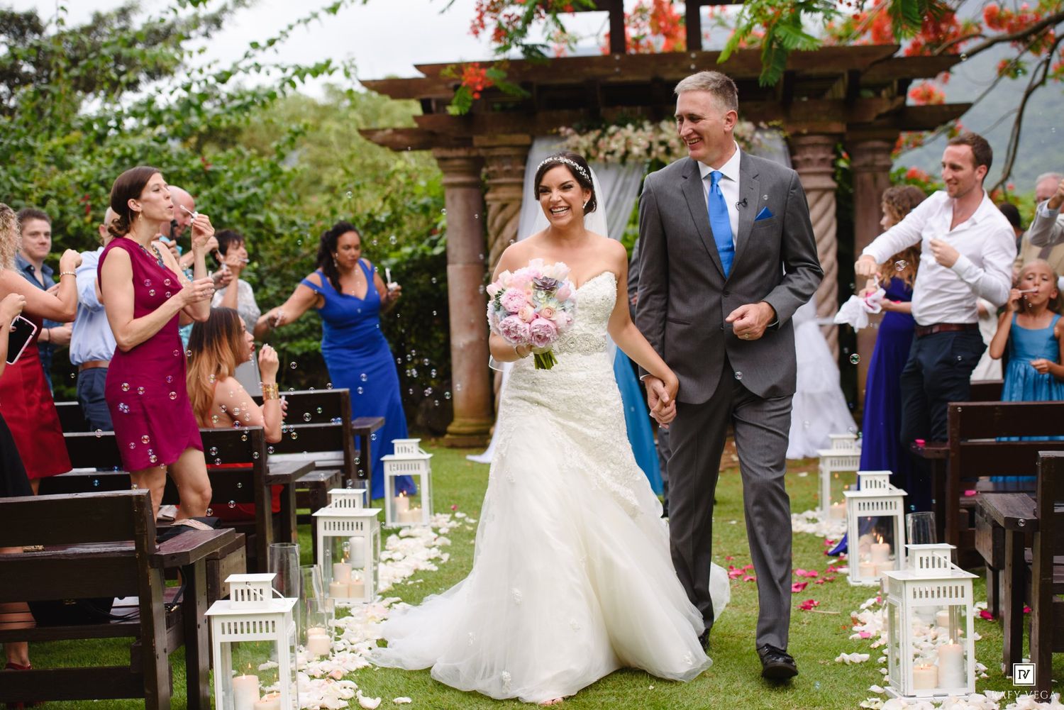 Bride and groom walk down the aisle at an outdoor wedding ceremony with guests celebrating and lanterns lining the path.