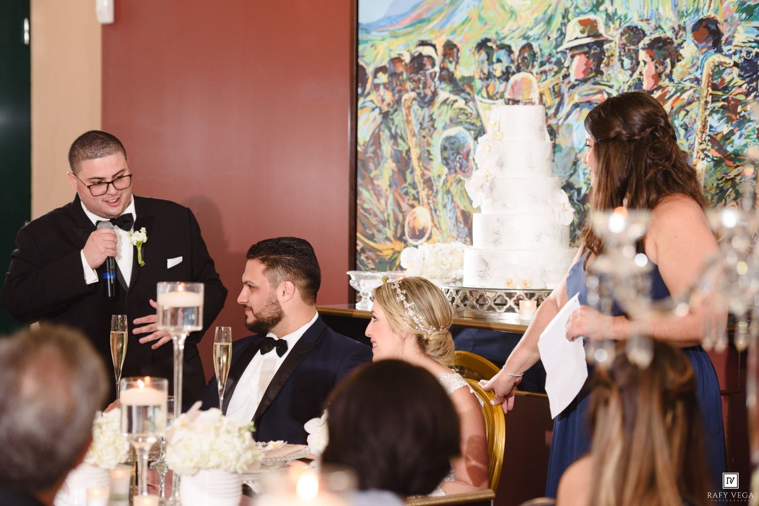 Wedding guests laugh during a speech at an elegant reception with a white tiered cake and colorful mural backdrop.