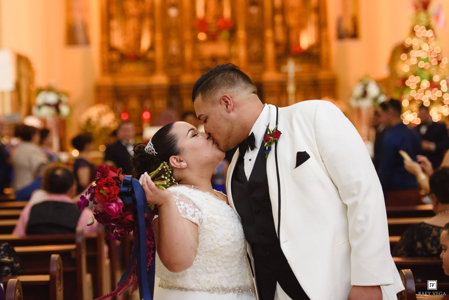 Bride and groom sharing first kiss at church altar during wedding ceremony with Christmas tree in background.