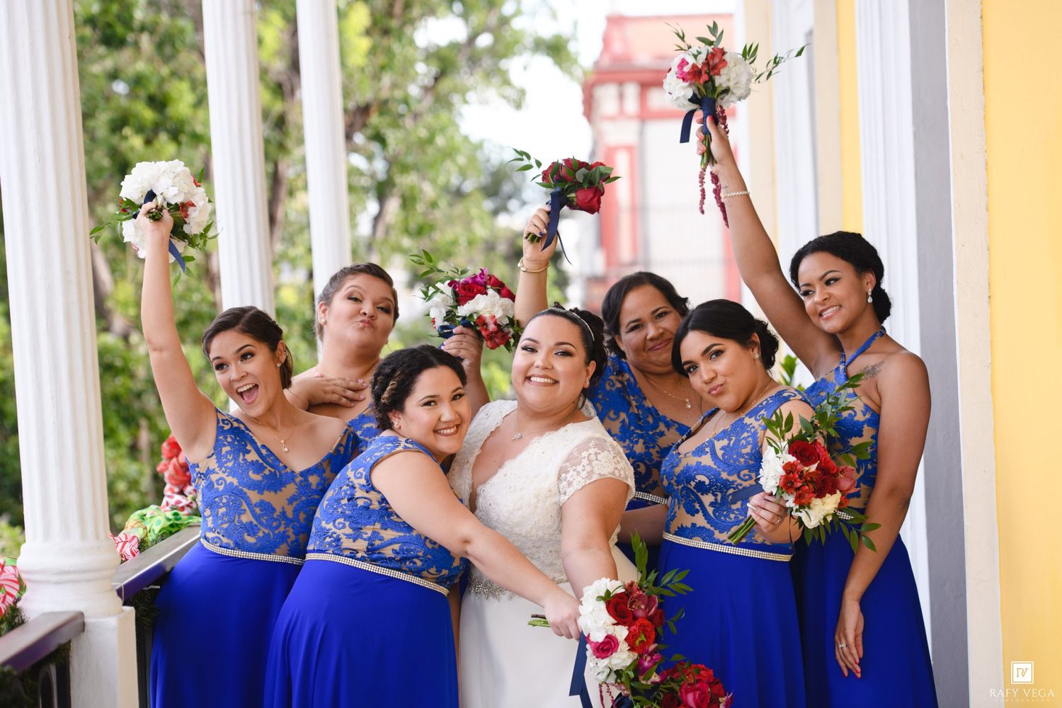 Bride in white gown poses with six bridesmaids in royal blue dresses holding bouquets aloft on a decorated porch.