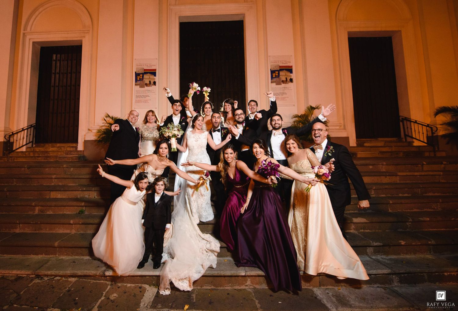Wedding party celebrating on church steps at night, bride in white gown with bridesmaids in purple and champagne dresses.