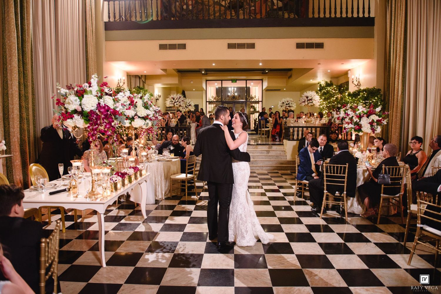 Bride and groom share first dance on checkered ballroom floor surrounded by guests and lush floral arrangements.