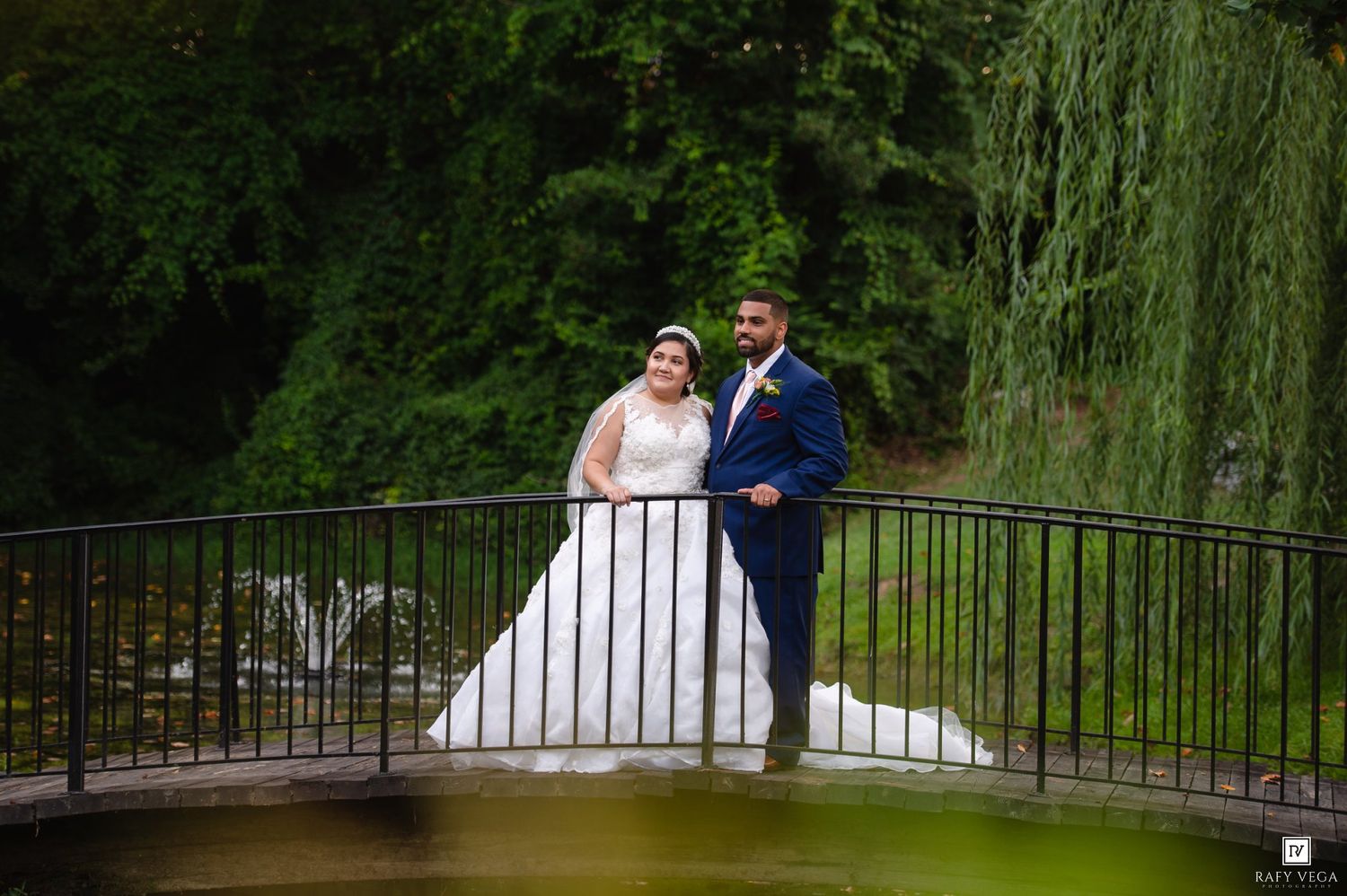 Bride and groom posing on a bridge surrounded by lush greenery and a willow tree on their wedding day.