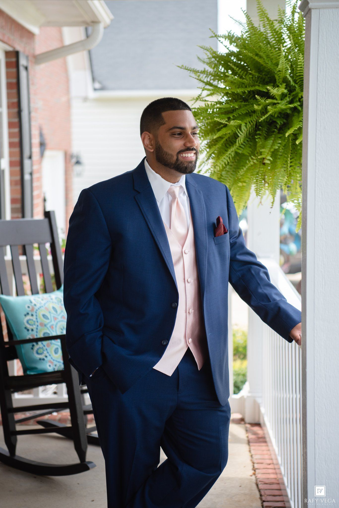 Smiling groom in navy blue suit with blush vest and pocket square posing on porch with fern backdrop.