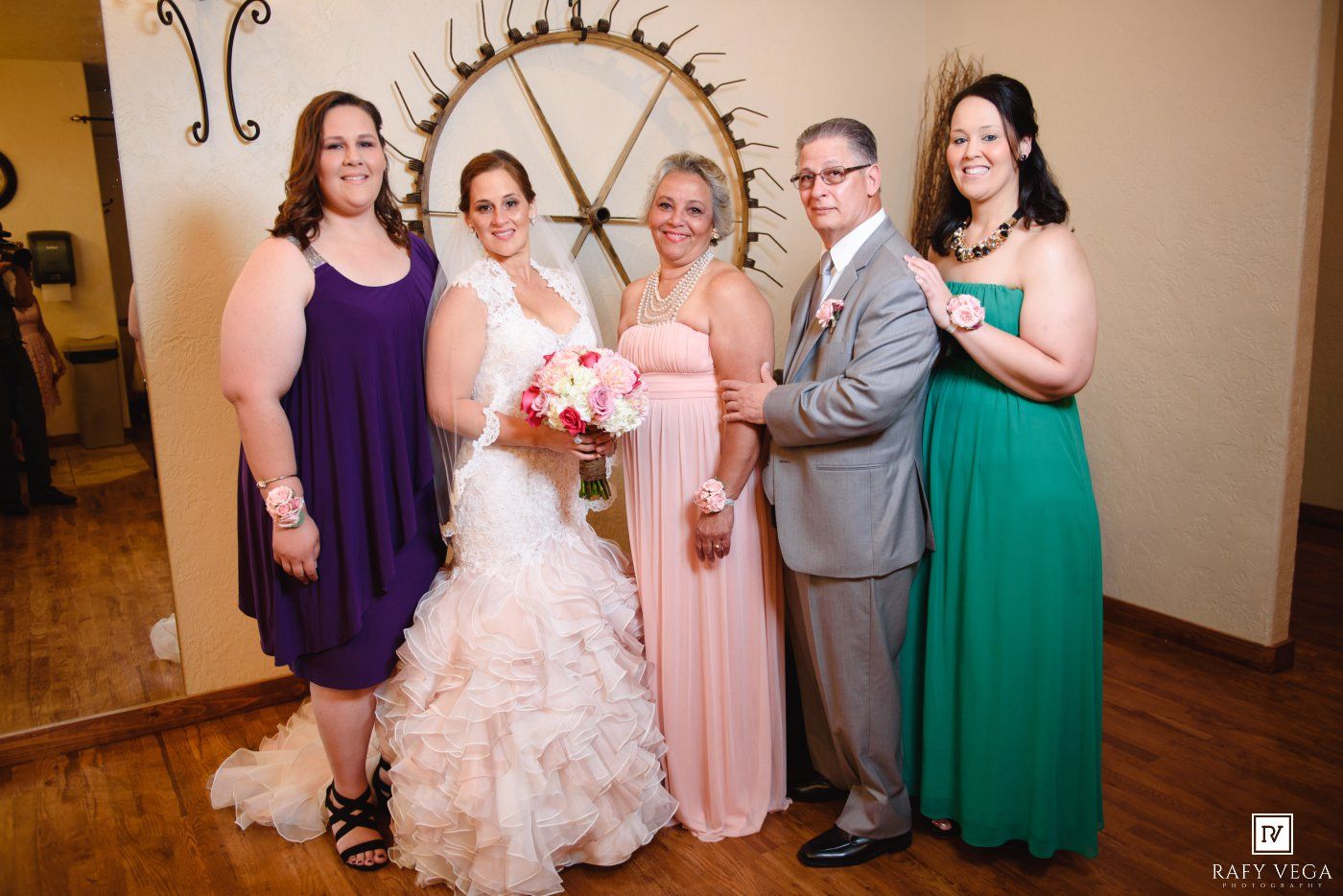 Wedding party group photo with bride in white gown, bridesmaids in purple and green dresses, and father of the bride.