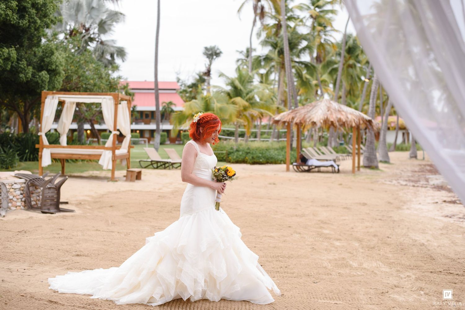 Red-haired bride in white ballgown wedding dress holding bouquet on sandy beach with tropical palm trees and ceremony arch.