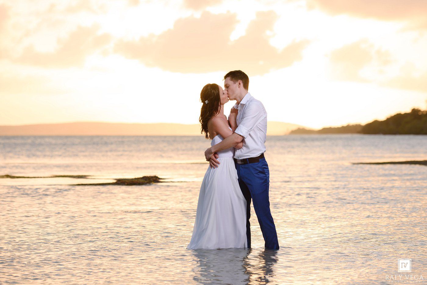 Newlyweds share a tender kiss standing in shallow ocean water during a golden tropical sunset.