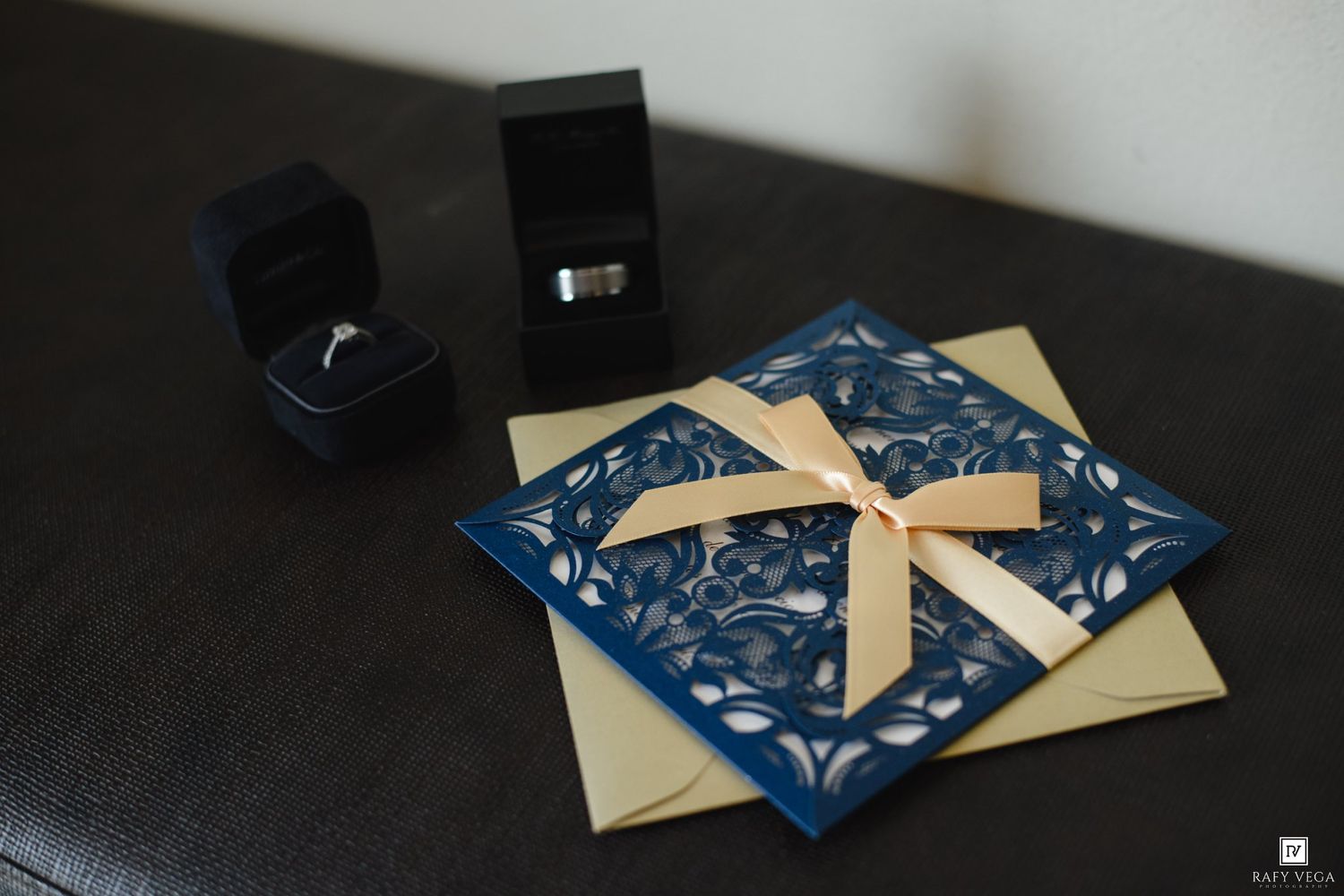 Elegant navy blue laser-cut wedding invitation with gold ribbon bow, beside two ring boxes on a dark surface.