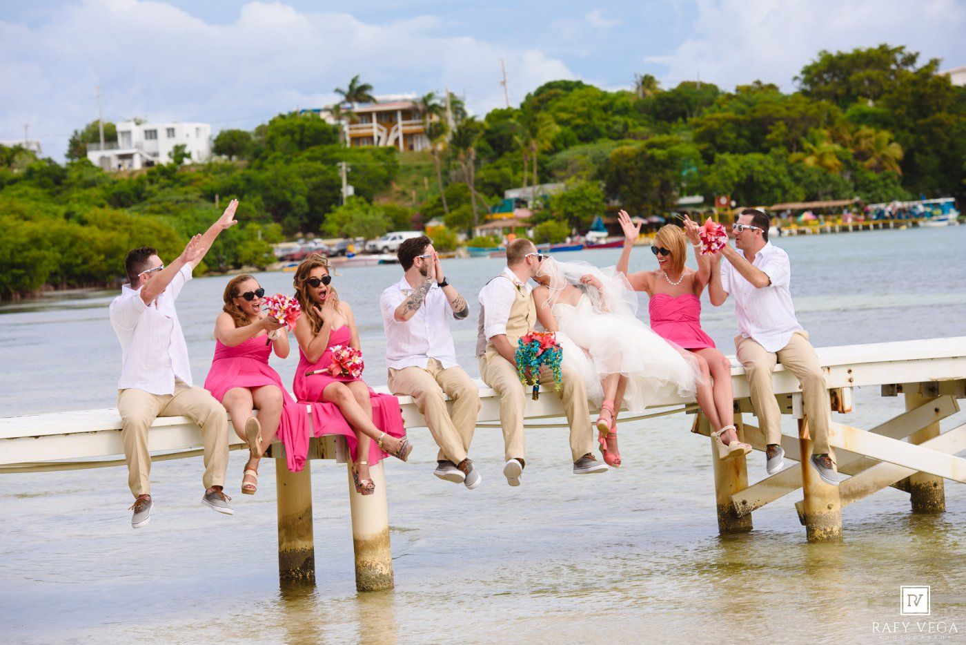 Wedding party in pink and white jumping off a dock over turquoise water at a tropical beach destination.