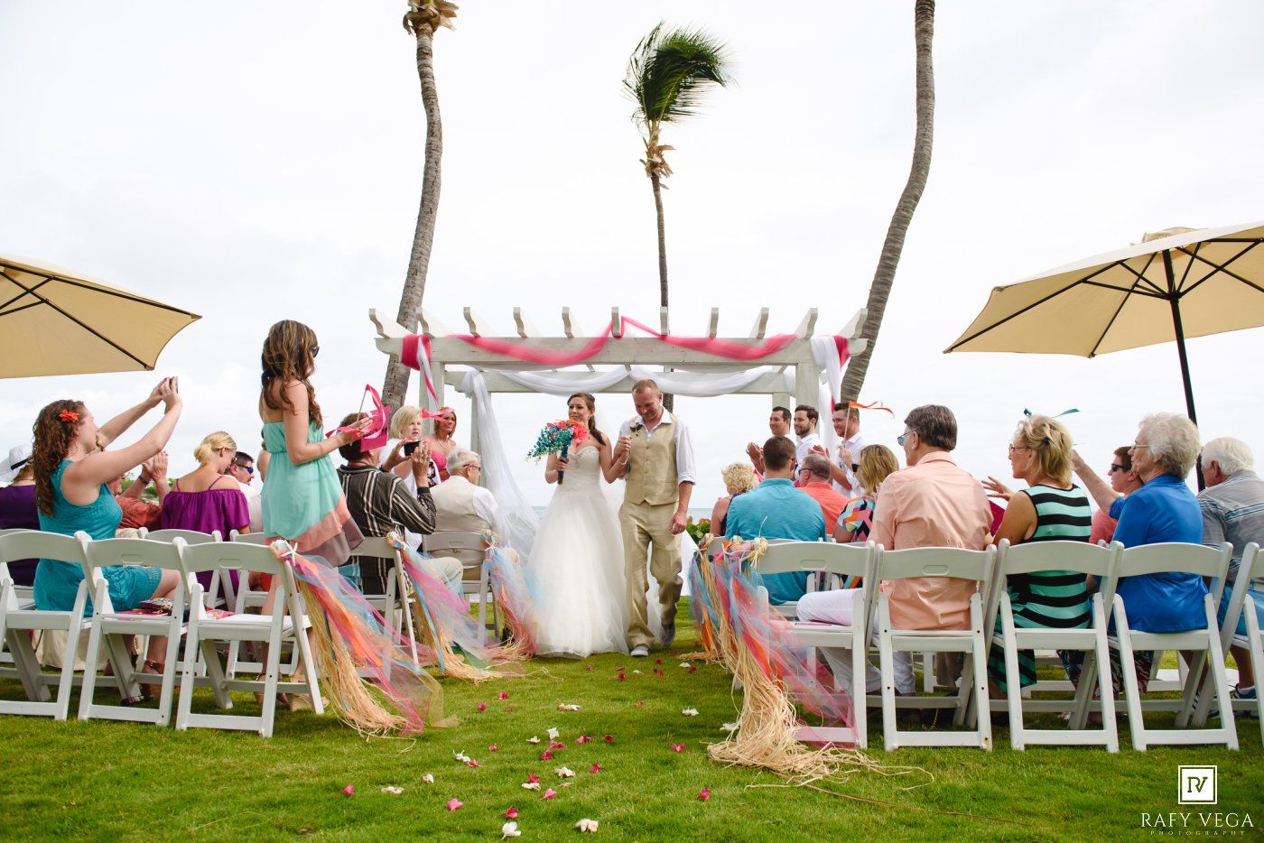 Outdoor beach wedding ceremony with bride and groom at floral arch, guests seated on white chairs under palm trees.