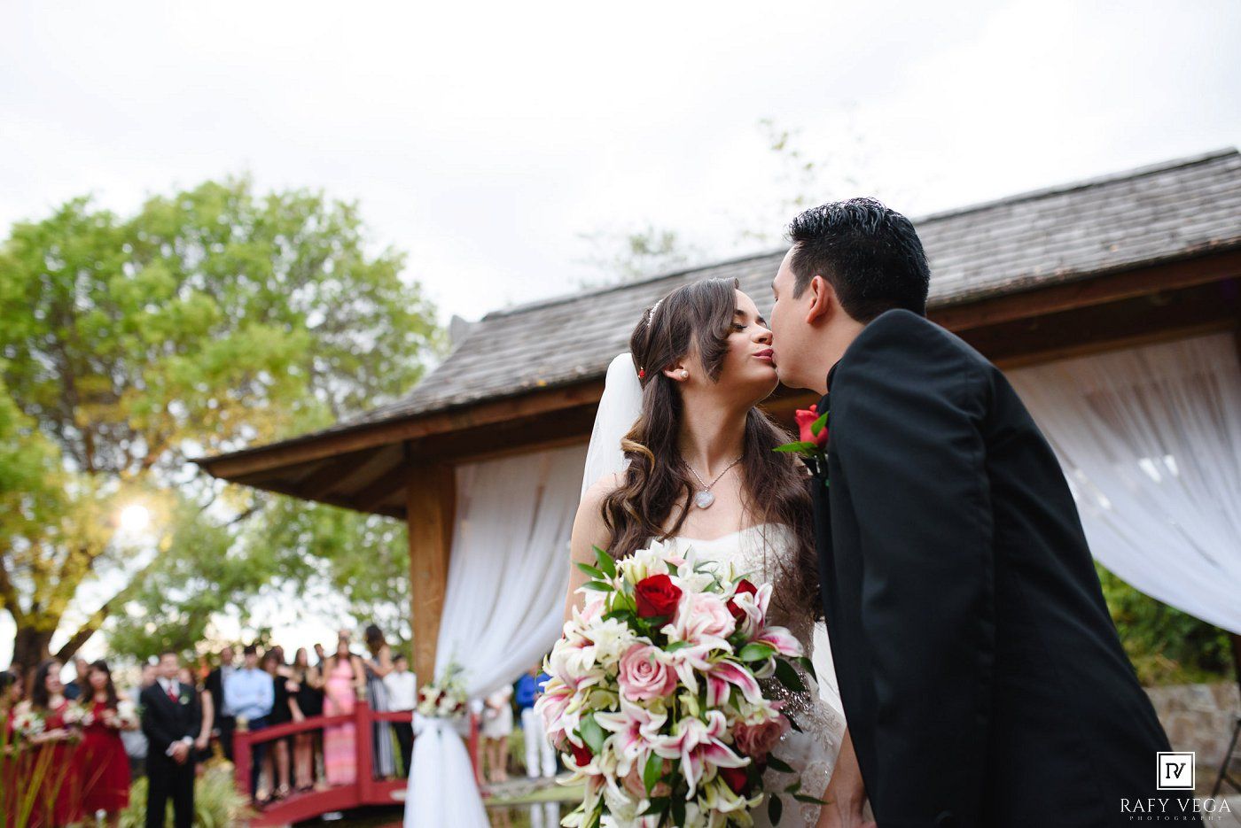 Newlywed couple shares a romantic kiss outdoors after their wedding ceremony with guests watching.