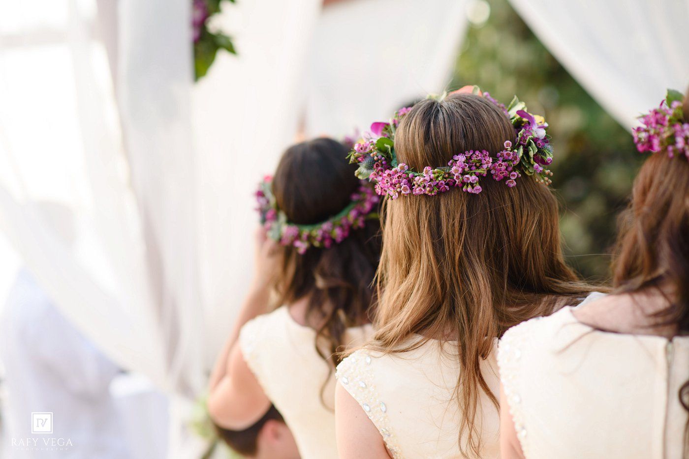 Bridesmaids wearing pink floral crowns stand together during an outdoor wedding ceremony.