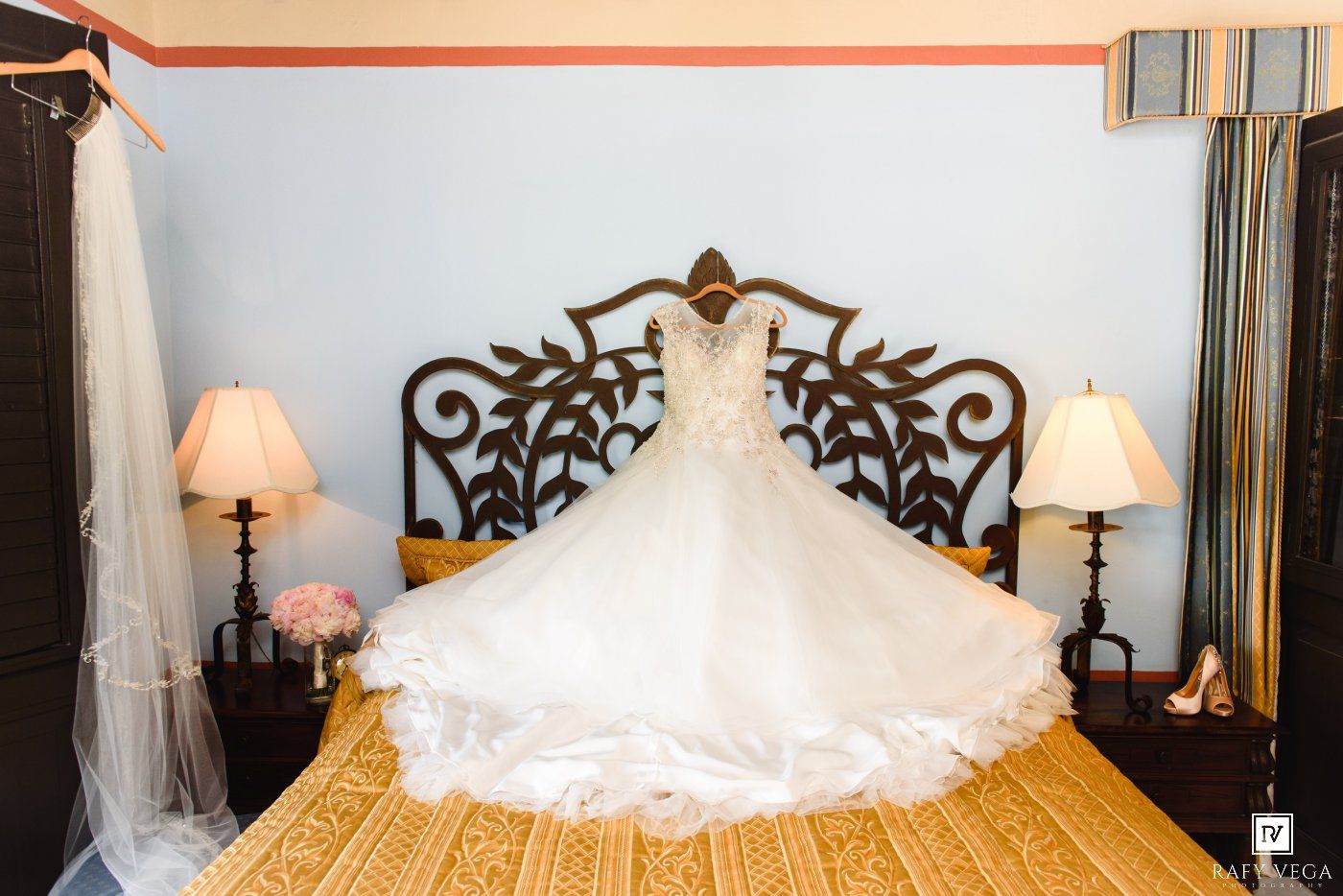 Elegant white wedding dress displayed on a bed with ornate headboard, flanked by lamps in a softly lit bridal suite.