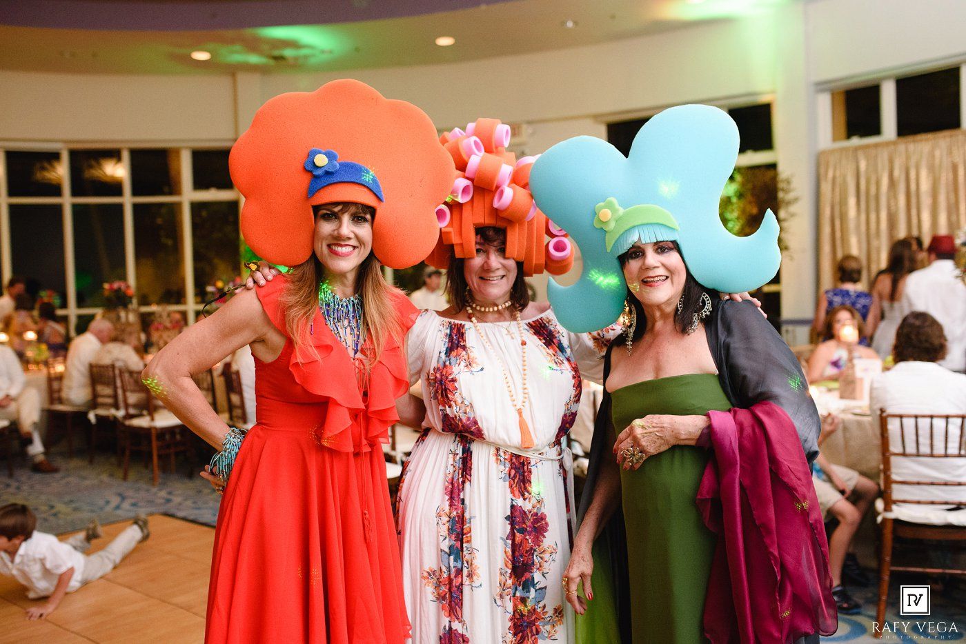 Three women in colorful floral hat costumes posing together at a festive indoor event.