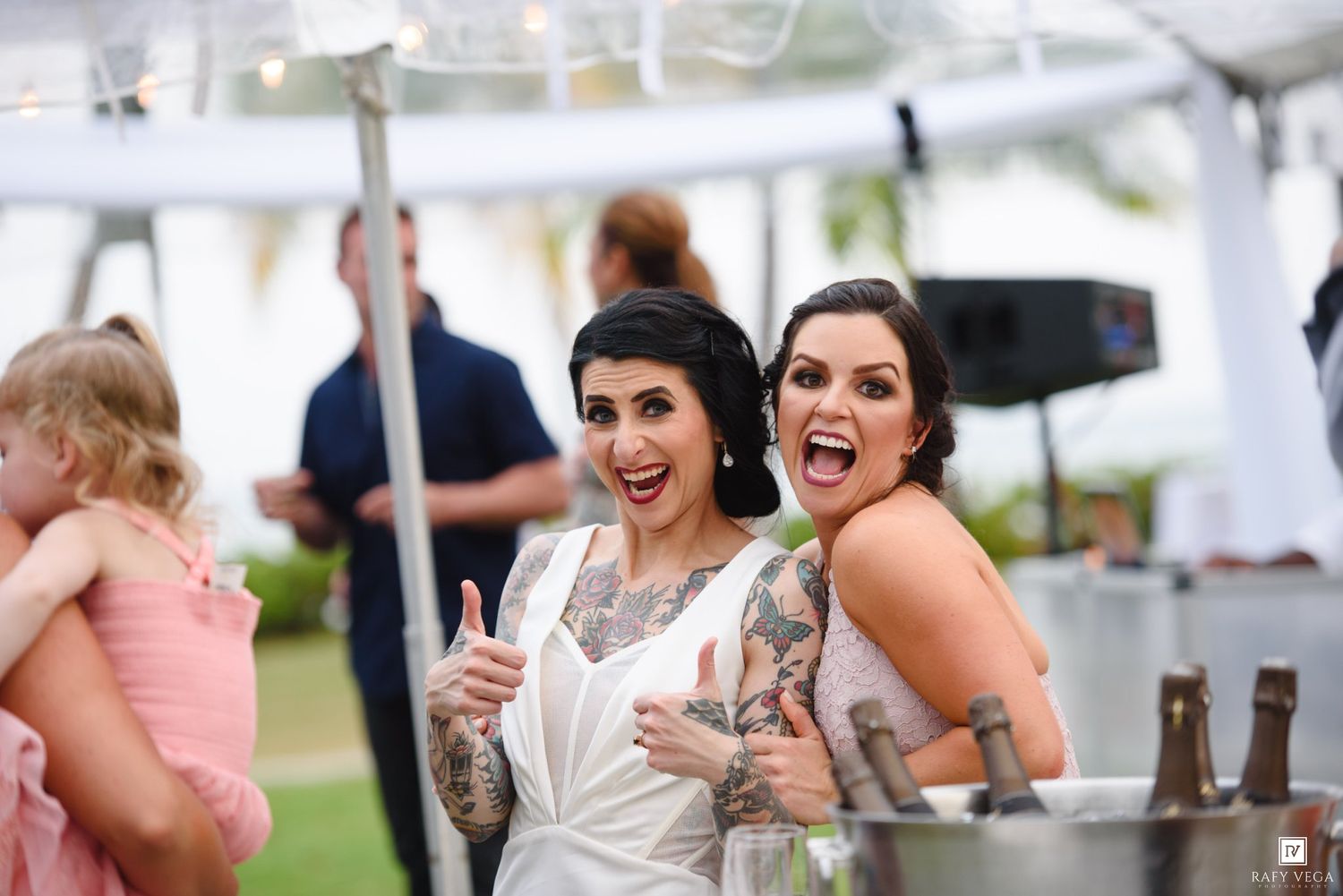 Two brides in wedding attire giving thumbs up and smiling at outdoor wedding reception under white tent.
