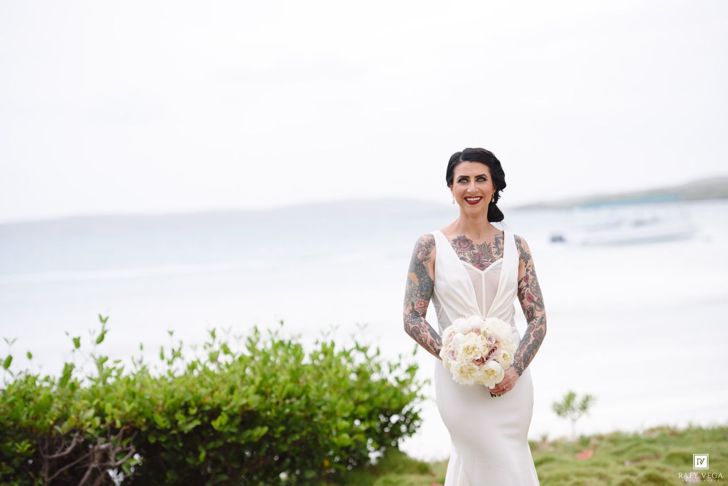 Beautiful tattooed bride in fitted white wedding dress holding white floral bouquet near seaside.