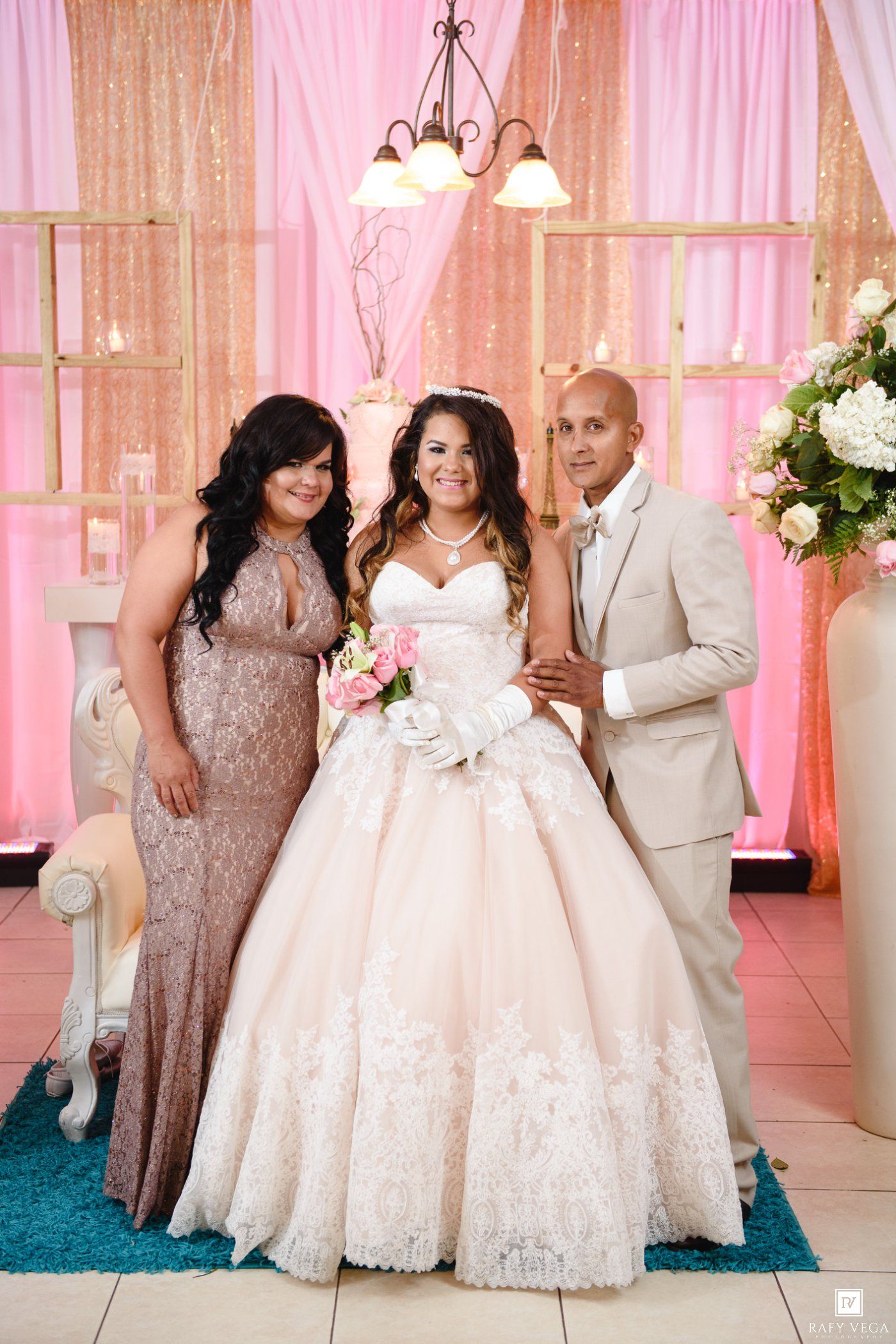 Bride in white ballgown poses with two guests at elegant pink-themed wedding reception venue.