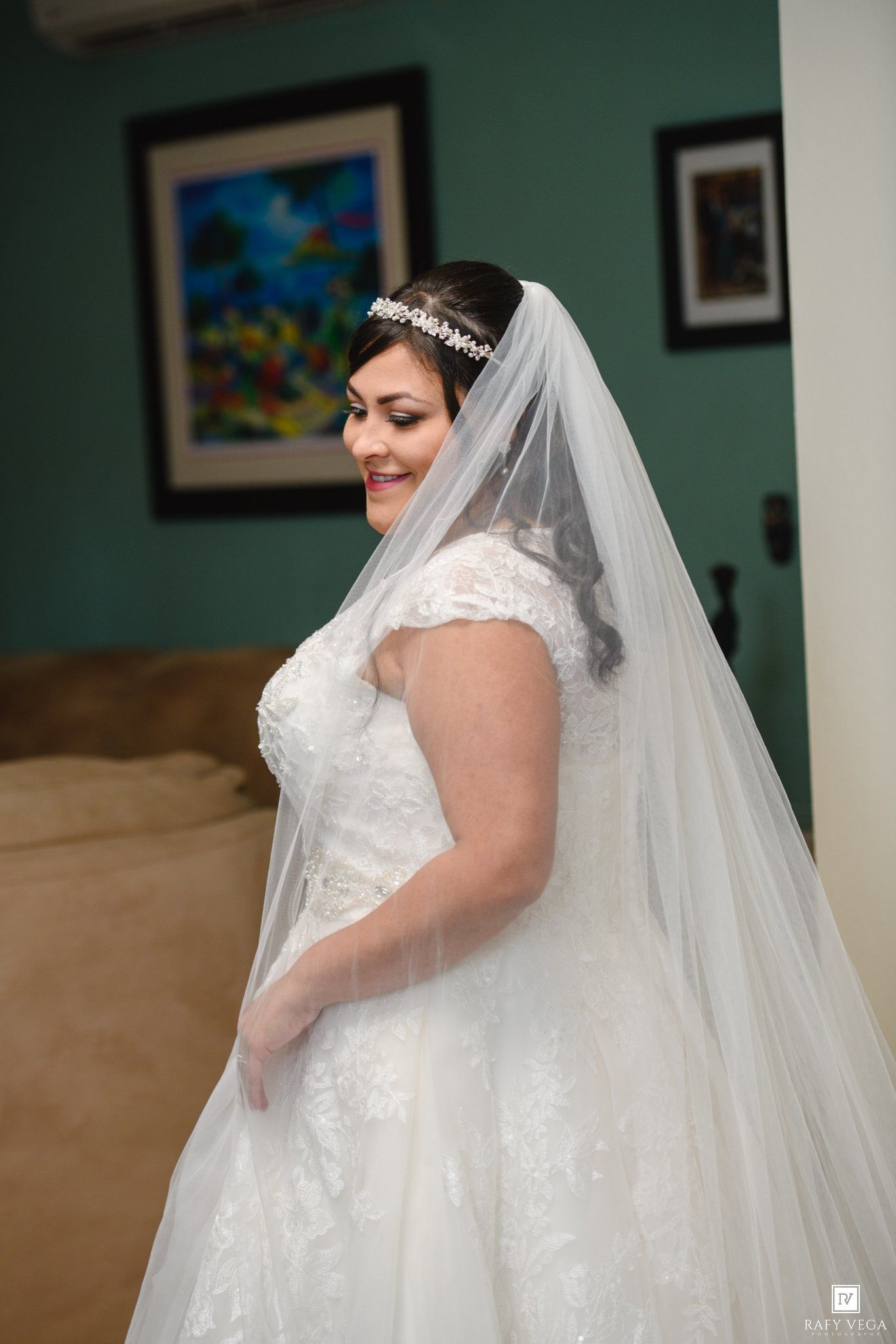 Bride in lace wedding gown with cathedral veil and crystal headpiece smiling in bridal suite.