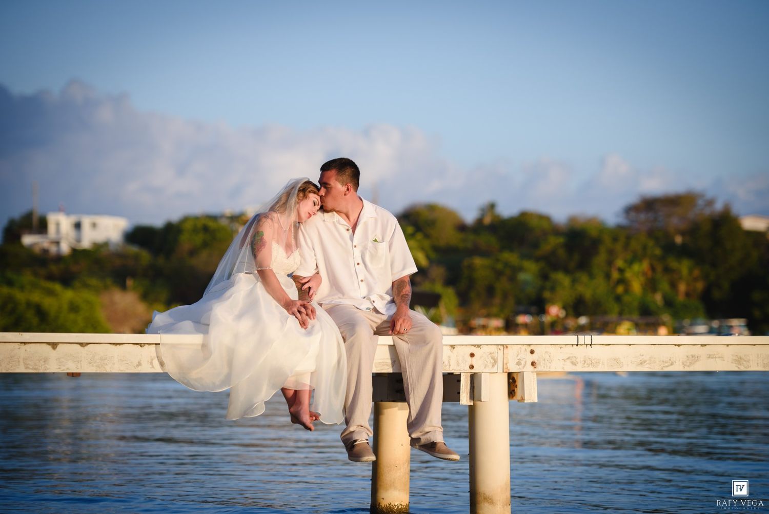 A bride and groom share a romantic kiss while sitting on a sunlit pier overlooking calm tropical waters.
