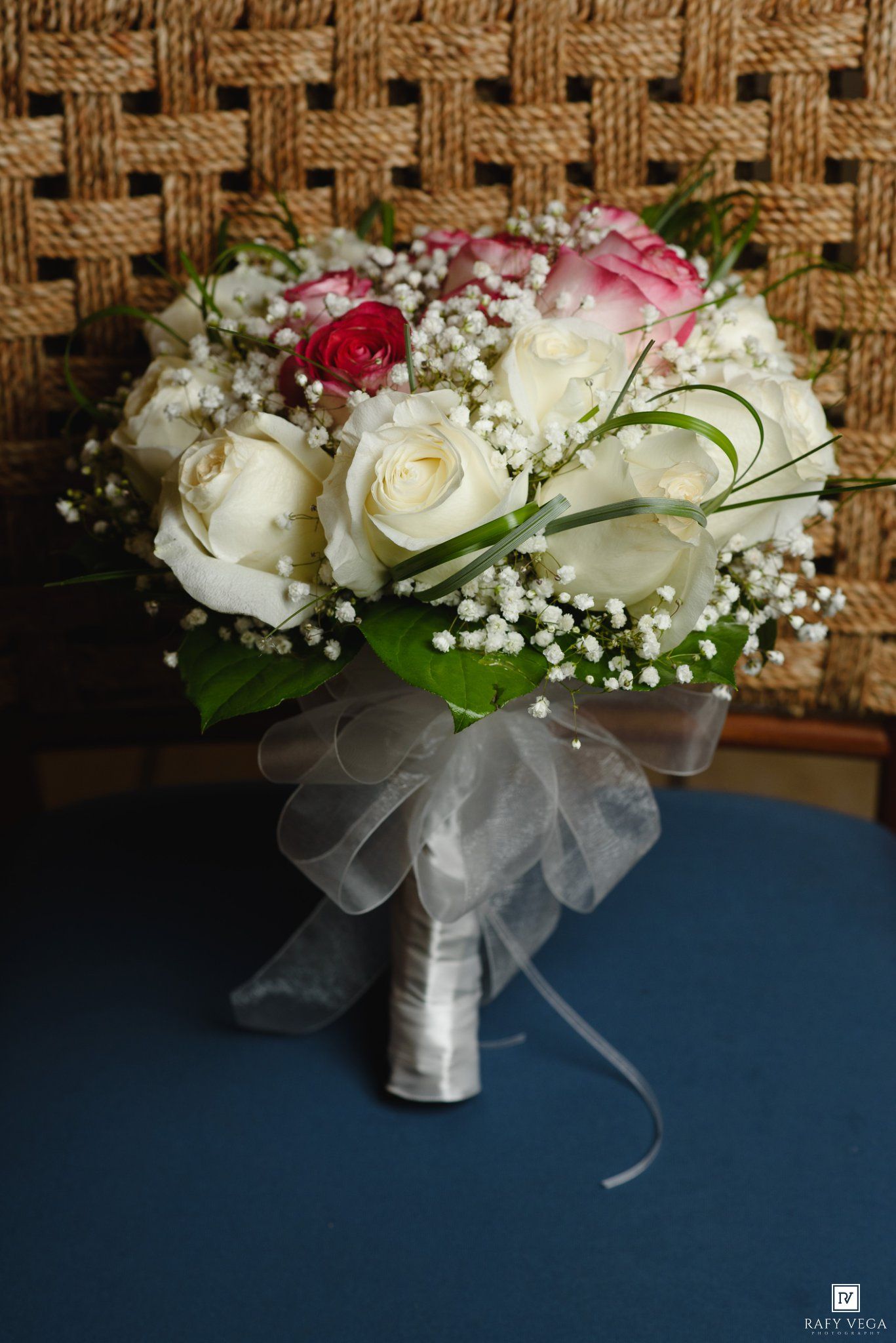 Elegant bridal bouquet featuring white and pink roses with baby's breath, tied with a silver ribbon, resting on a blue surface.