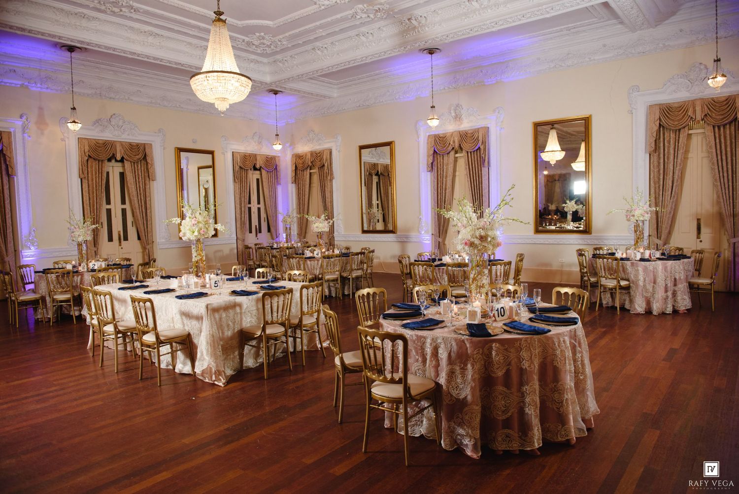 Luxurious ballroom wedding reception with gold chiavari chairs, navy blue linens, and tall floral centerpieces.