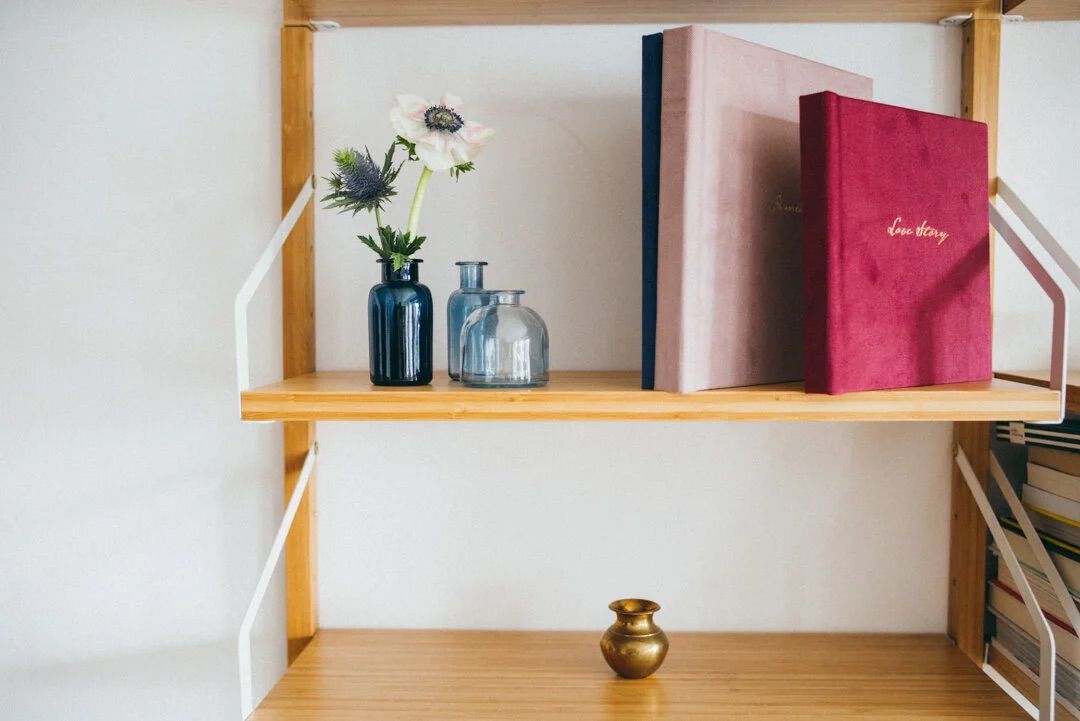 Wooden shelf with pink and blue books, small glass vases with flowers, and a gold brass pot decoration.