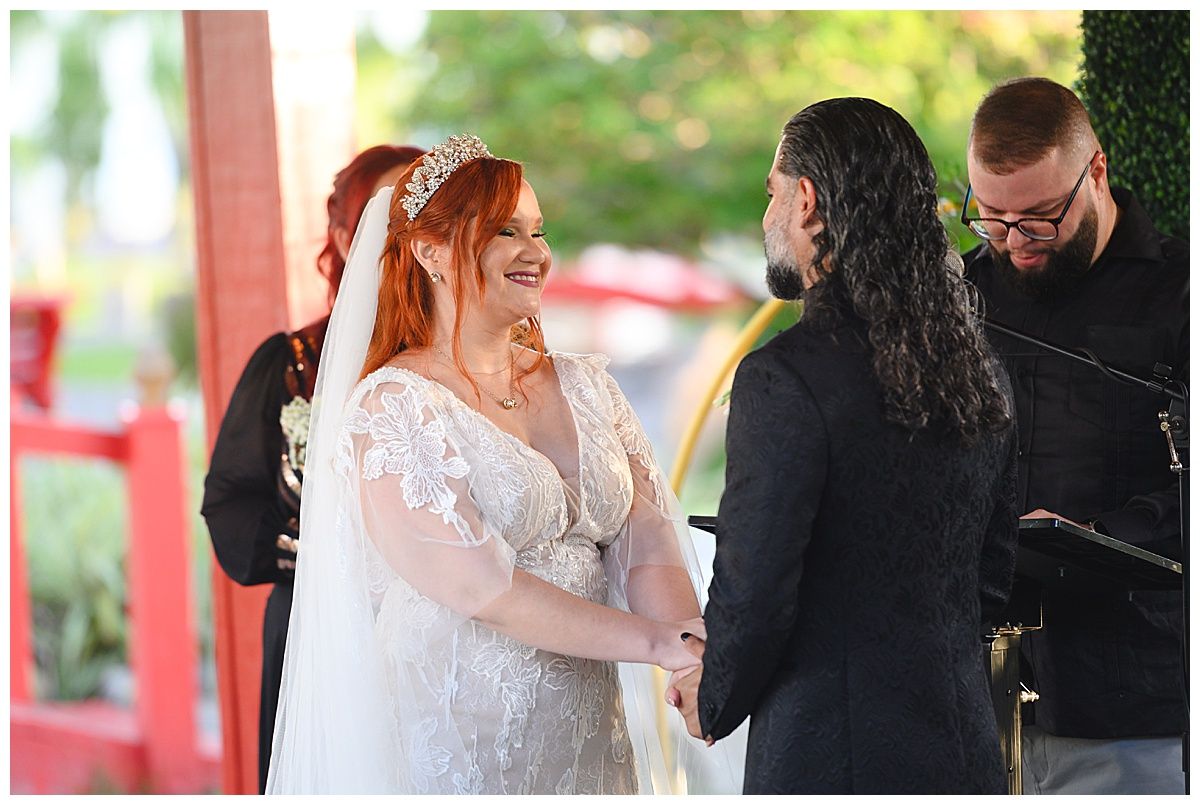 Bride in lace wedding gown and tiara smiles at groom during outdoor wedding ceremony with officiant.