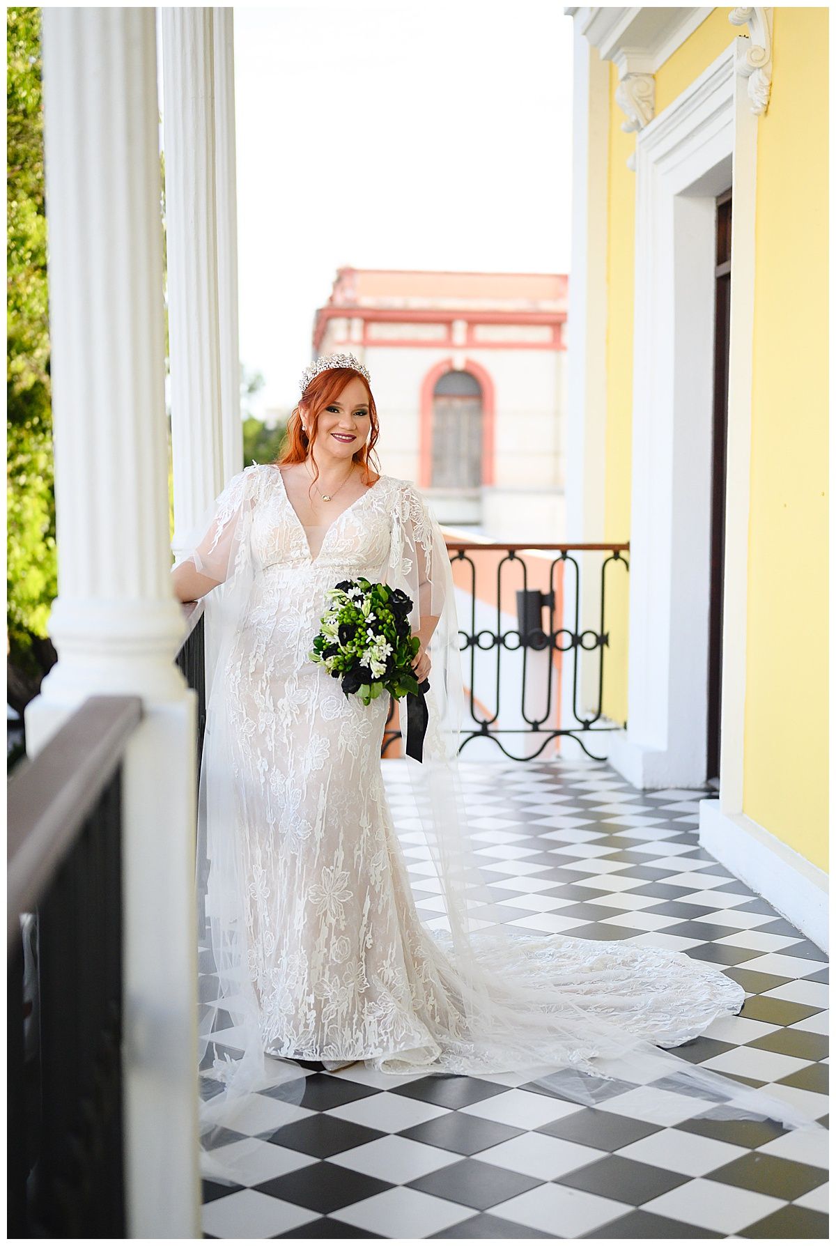 Bride in lace wedding gown holding green bouquet on checkered balcony of colorful building in Puerto Rico.