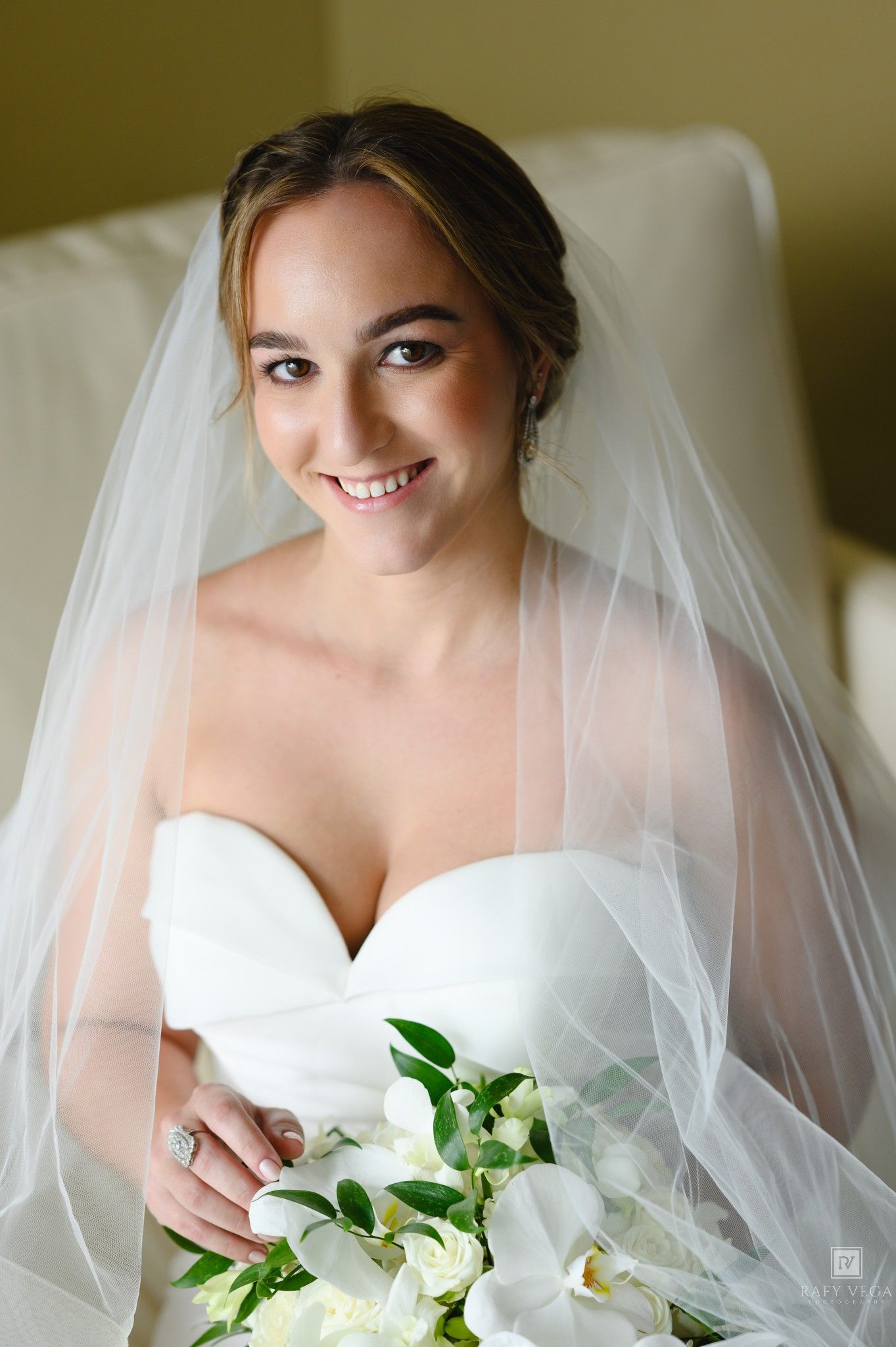 Smiling bride in white strapless gown and veil holds white orchid bouquet while seated, showcasing elegant bridal look.