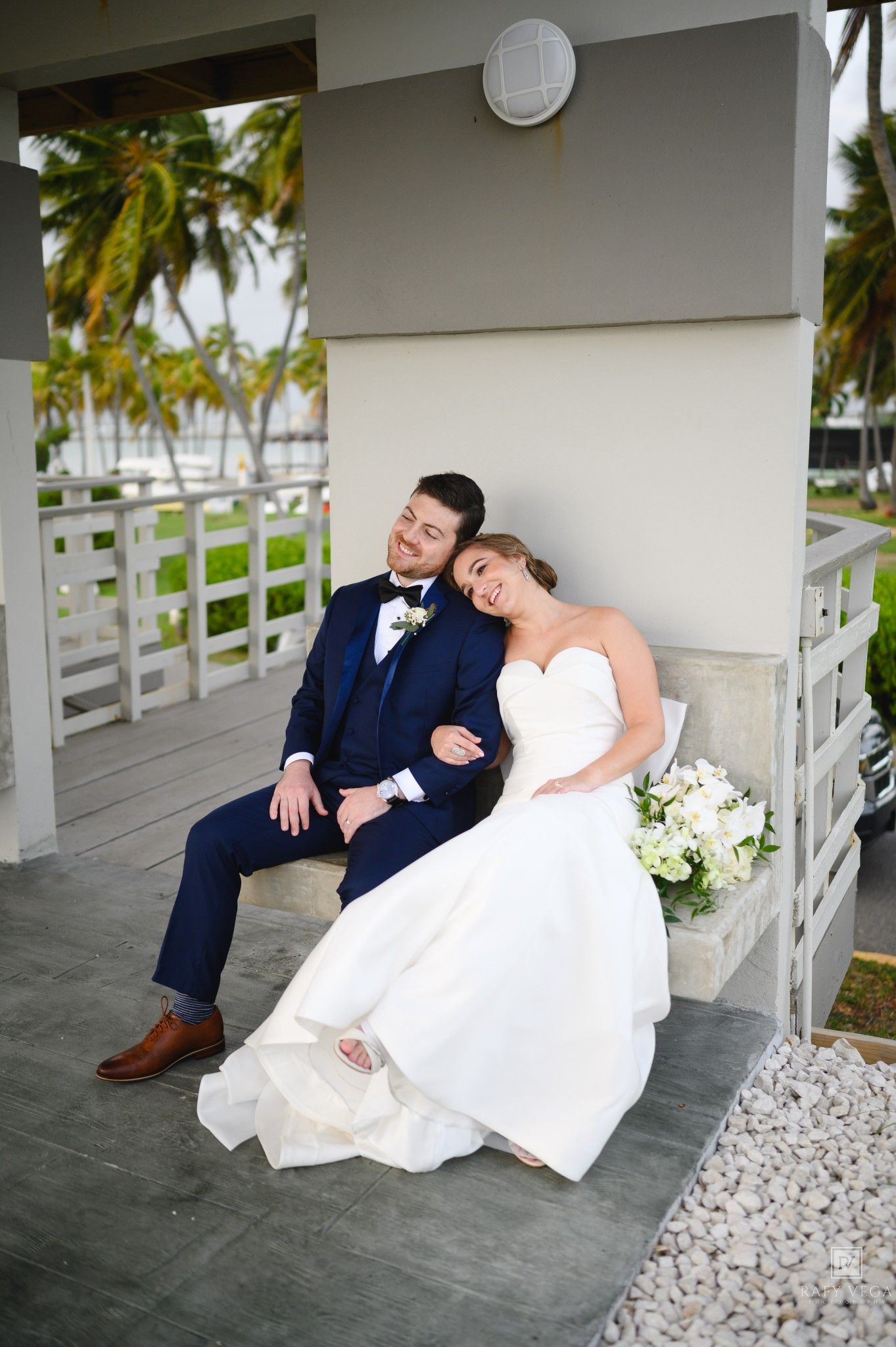 Newlyweds sit together on a porch bench, bride in white gown leaning on groom in navy suit, tropical venue backdrop.