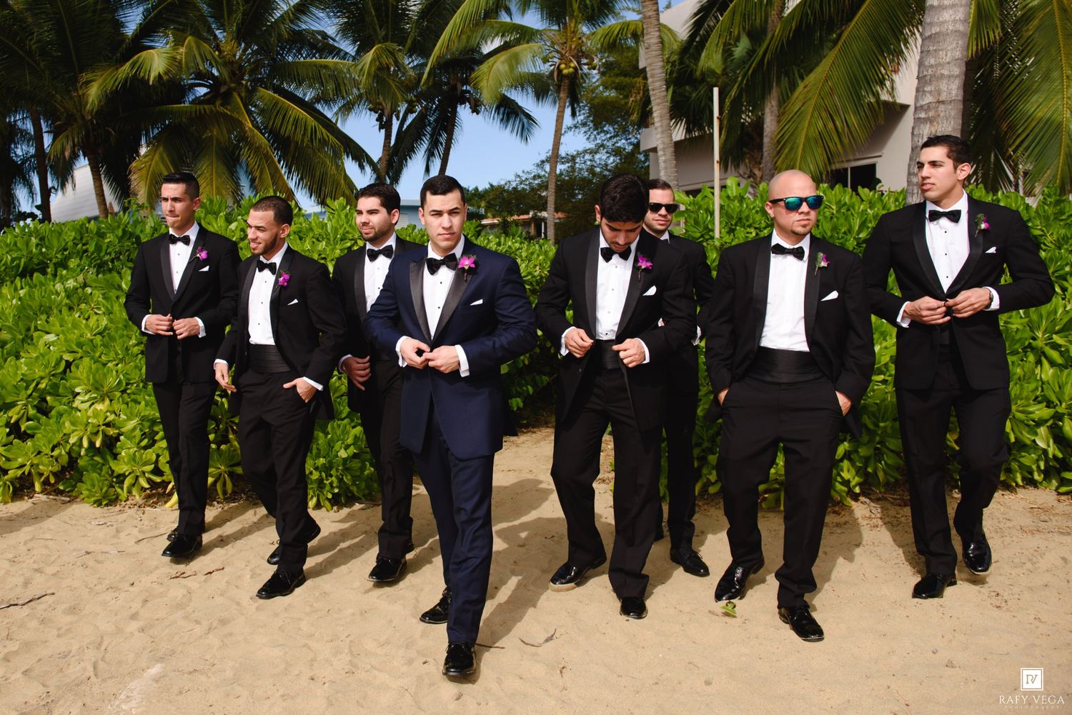 Groom and groomsmen in black tuxedos with bow ties and pink boutonnieres posing on a tropical beach.