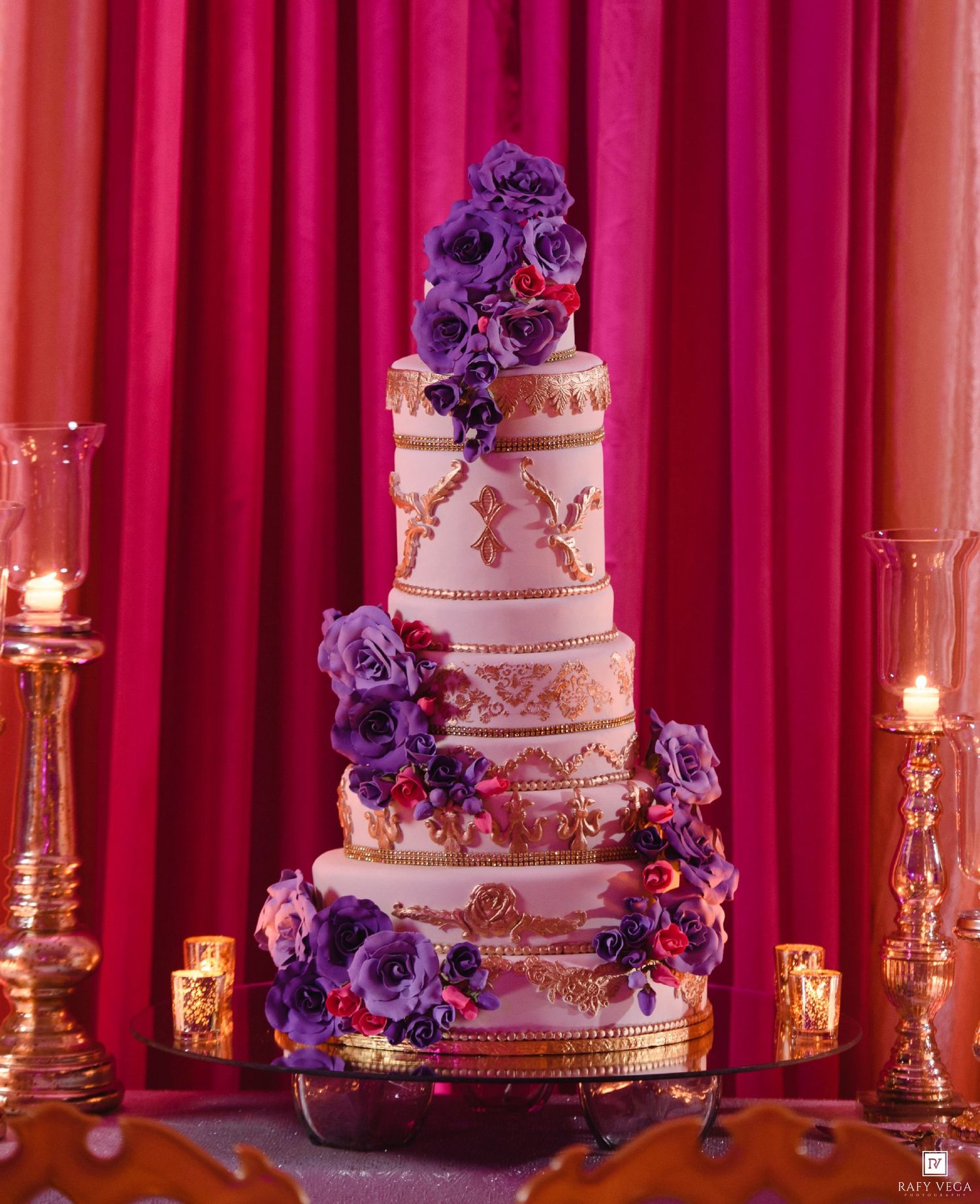 Elegant five-tier wedding cake with purple roses and gold detailing, flanked by candles against a pink backdrop.