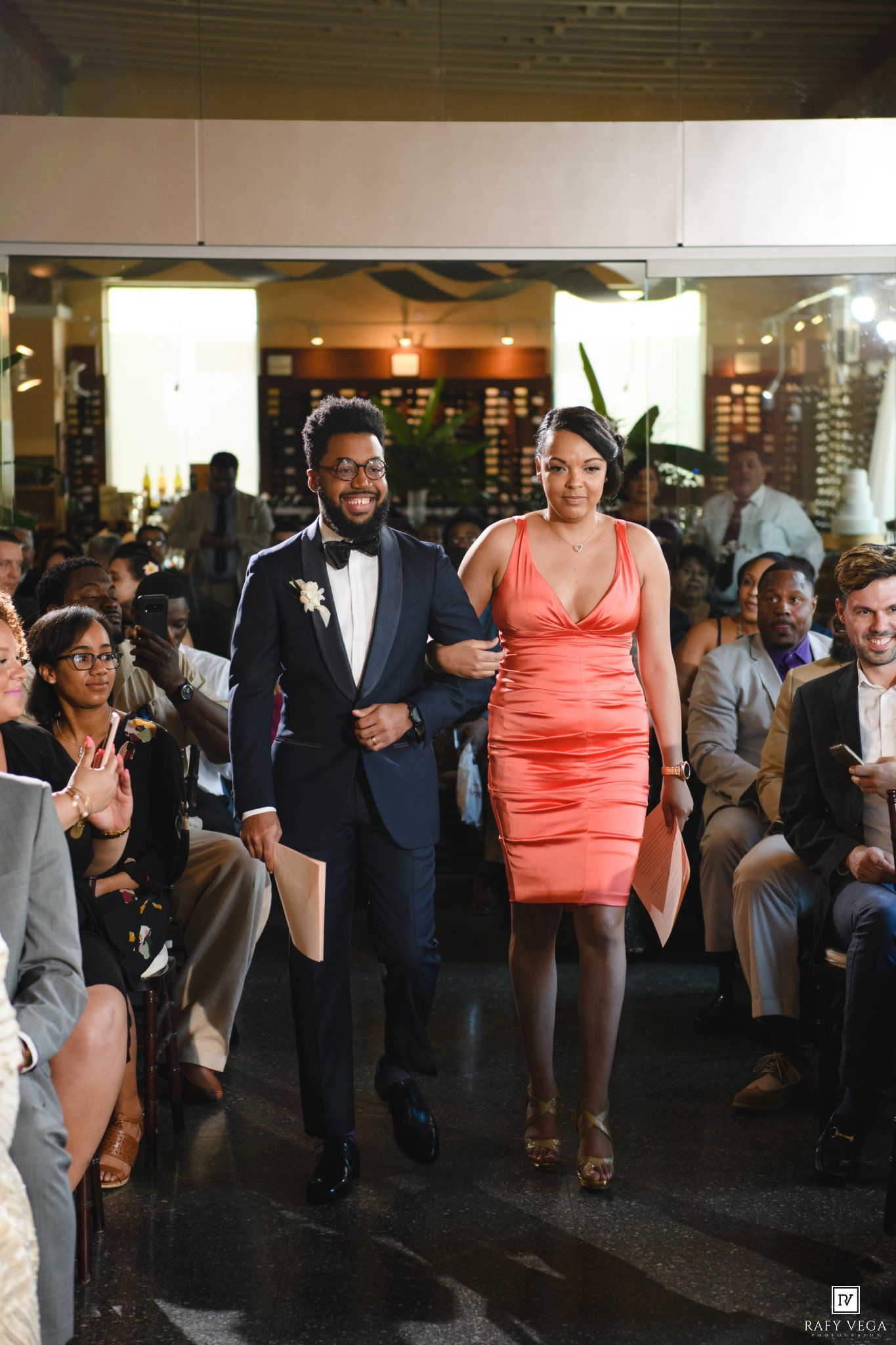 A groom in a tuxedo escorts a woman in a coral dress down the aisle at a wedding reception.