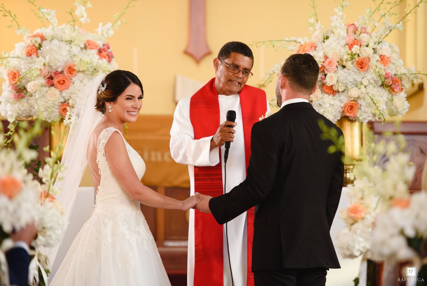 Bride and groom hold hands as priest officiates wedding ceremony flanked by white and peach floral arrangements.