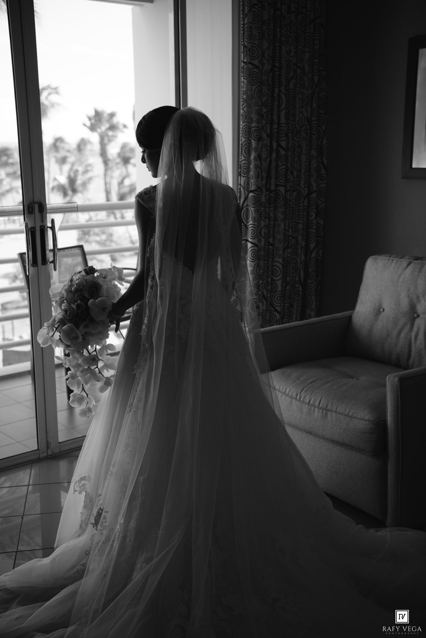 Black and white photo of a bride in a long veil and gown holding a bouquet, standing by a sunlit balcony door.