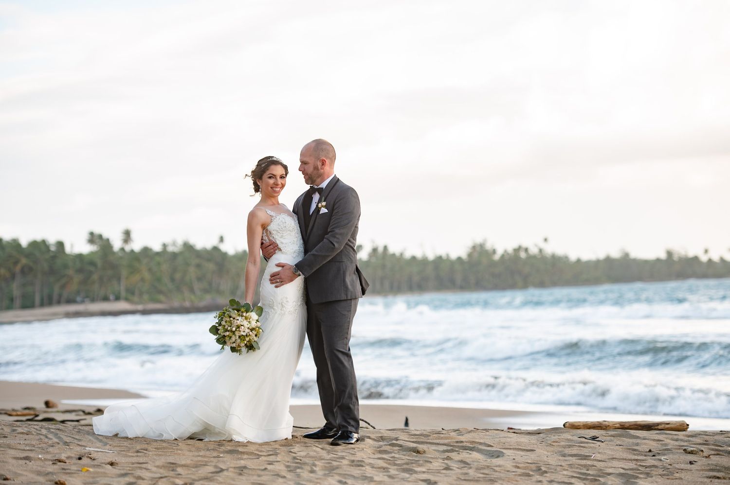 Bride and groom embracing on a tropical beach with ocean waves and palm trees in the background.
