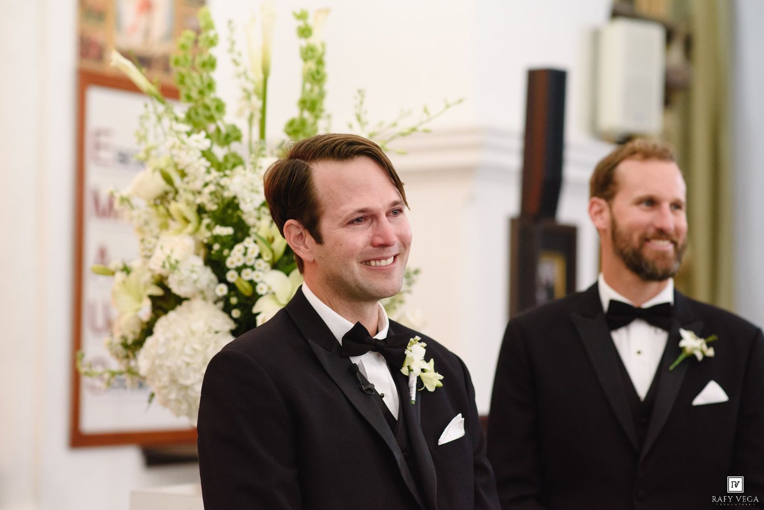 Groom in tuxedo smiles joyfully at altar with groomsman beside floral arrangement.