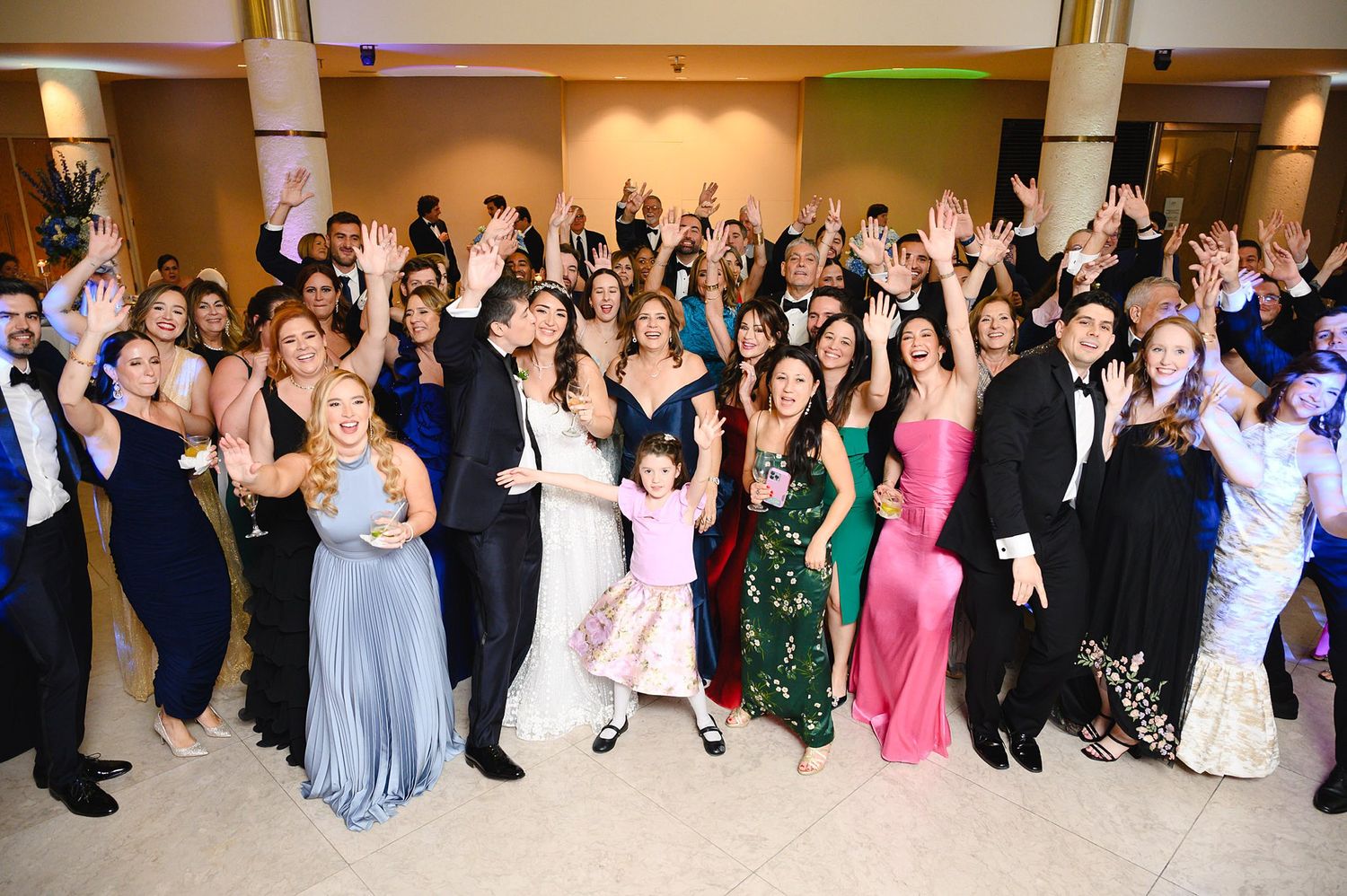 Large group of wedding guests in formal attire smiling and waving at a festive wedding reception.