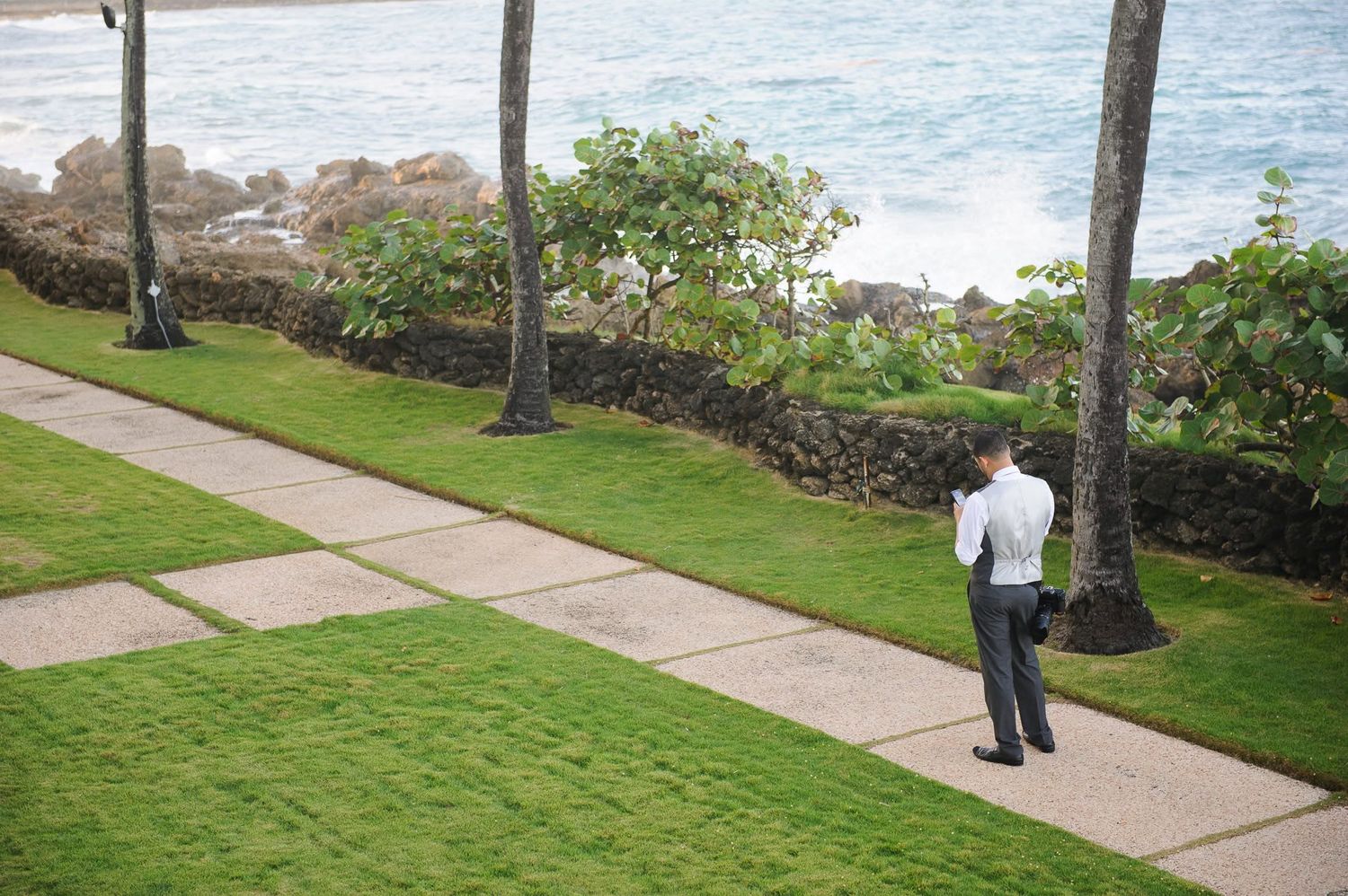 Man in white shirt walking along a lush green lawn pathway near a tropical ocean coastline with palm trees.