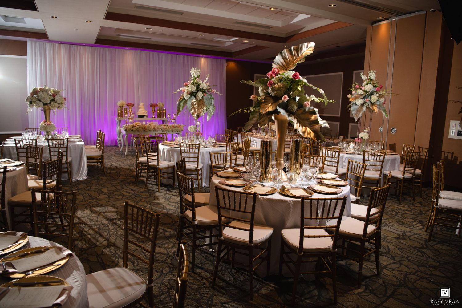 Elegant ballroom reception with gold chiavari chairs, tall tropical floral centerpieces, and purple uplighting on draped walls.