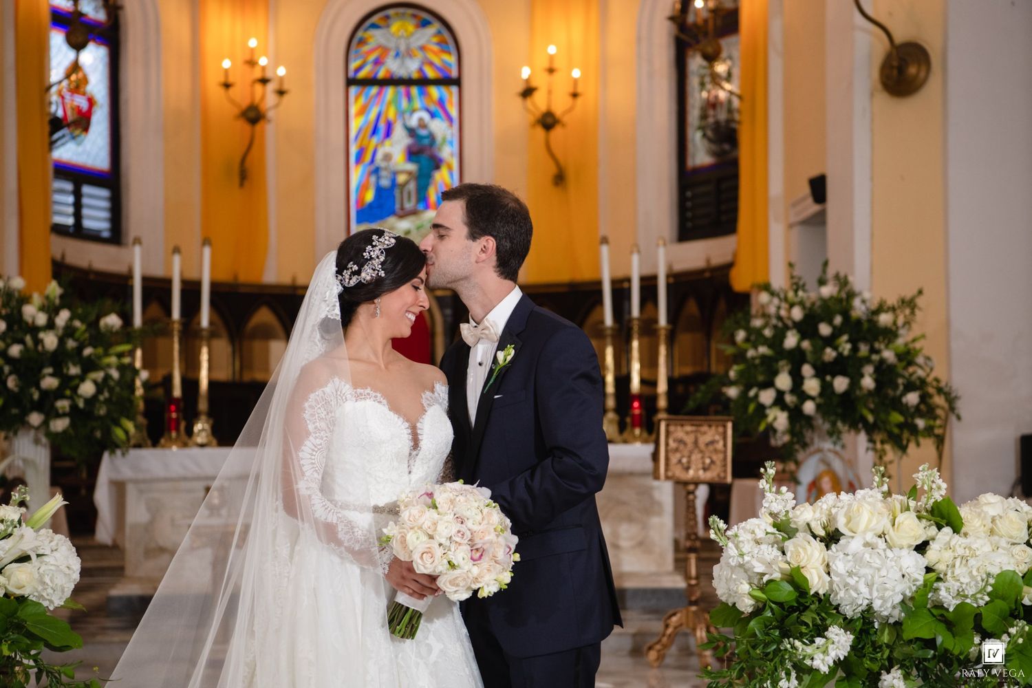 Bride and groom share a kiss at the altar of an elegant church adorned with white floral arrangements and candlelight.