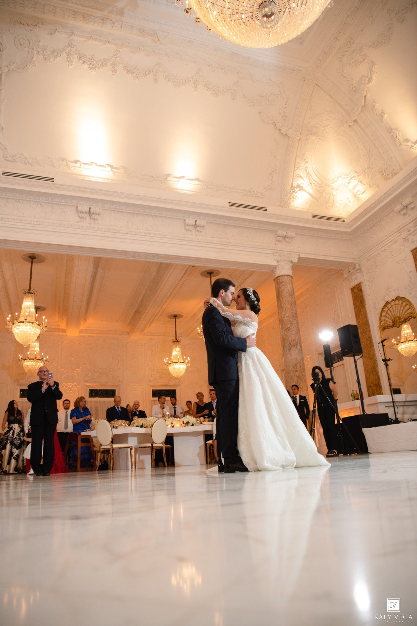 Bride and groom share their first dance in an elegant white ballroom with ornate ceilings and chandeliers at their wedding reception.